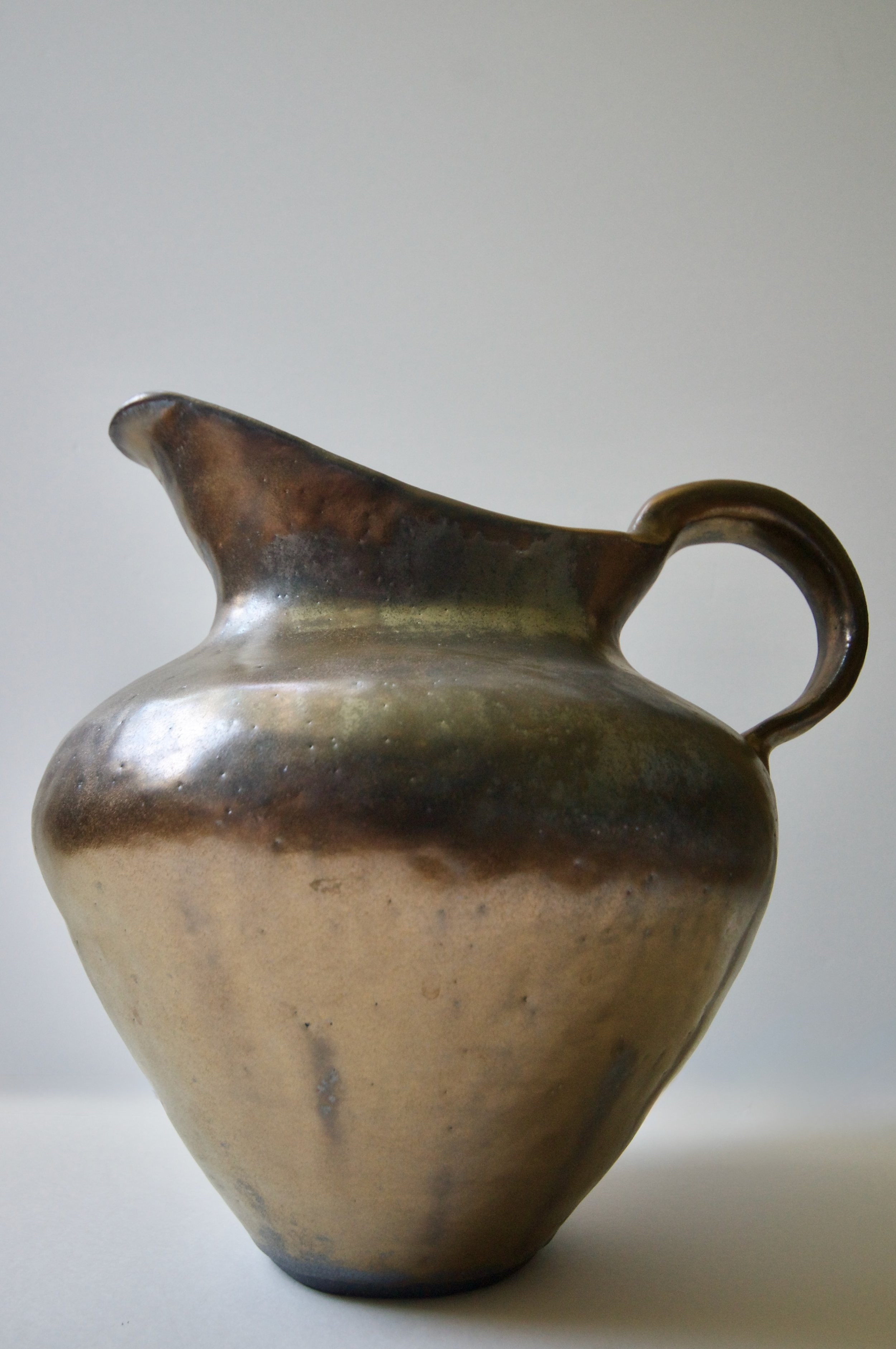 A ceramic vessel with a rounded body and a narrow neck, featuring a curved handle on the side, glazed in earthy tones of beige, brown, and hints of dark green.