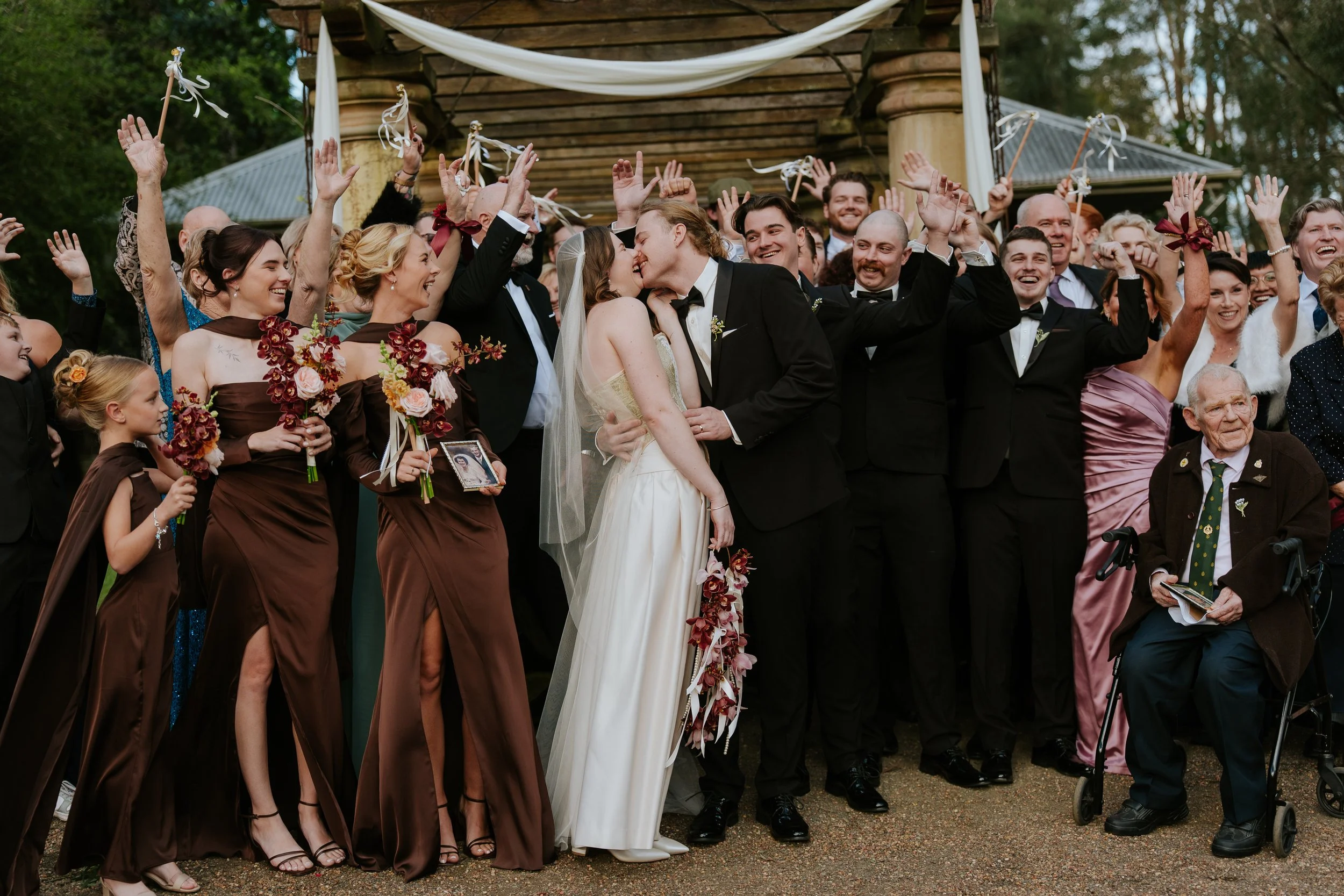 A group of people at a wedding celebration, with the bride and groom kissing in the center, surrounded by friends and family, some holding signs and bouquets, under a wooden pavilion outdoors.