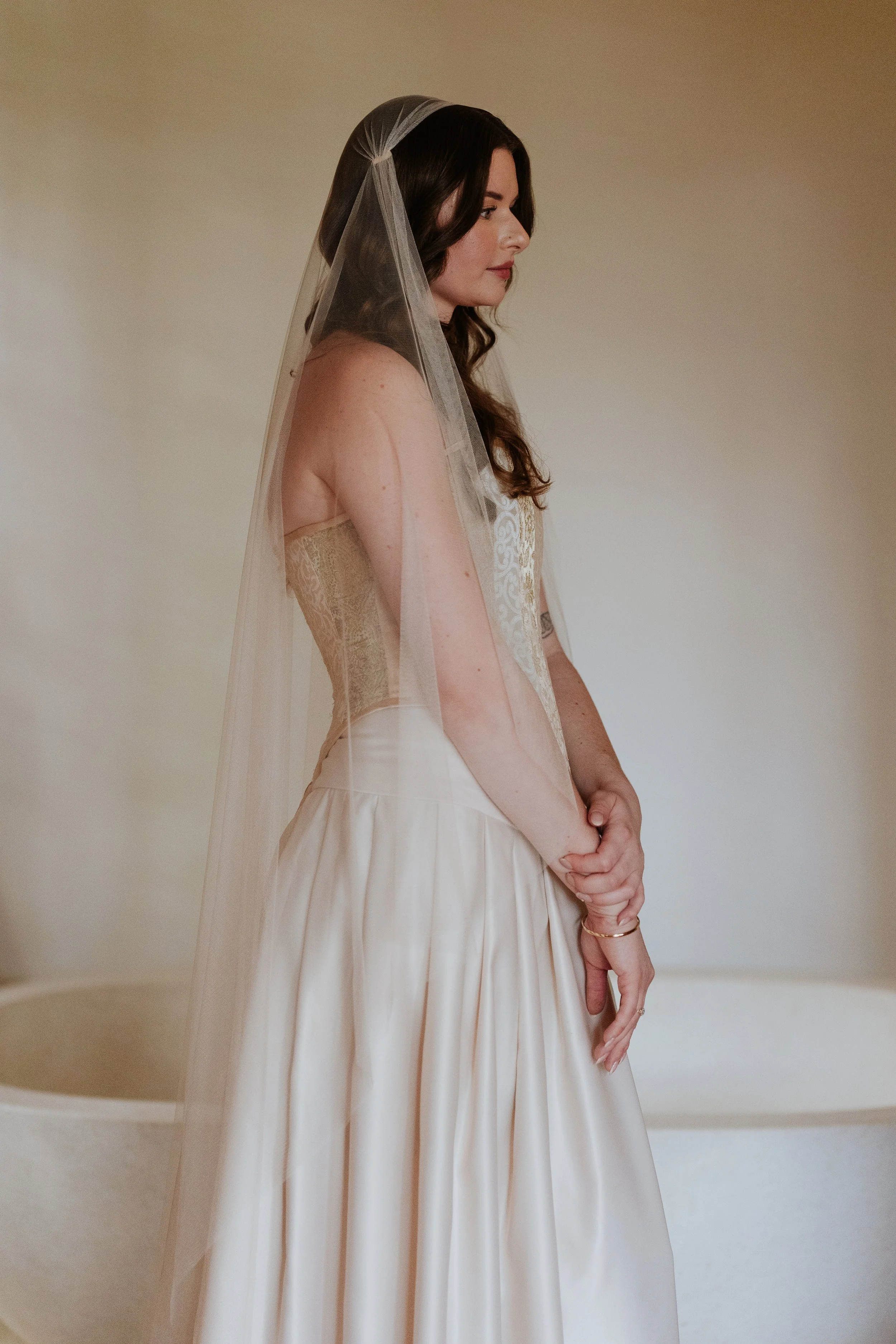 A woman dressed in a wedding gown with long brown hair and a sheer veil, standing sideways with her hands clasped, against a plain beige wall.
