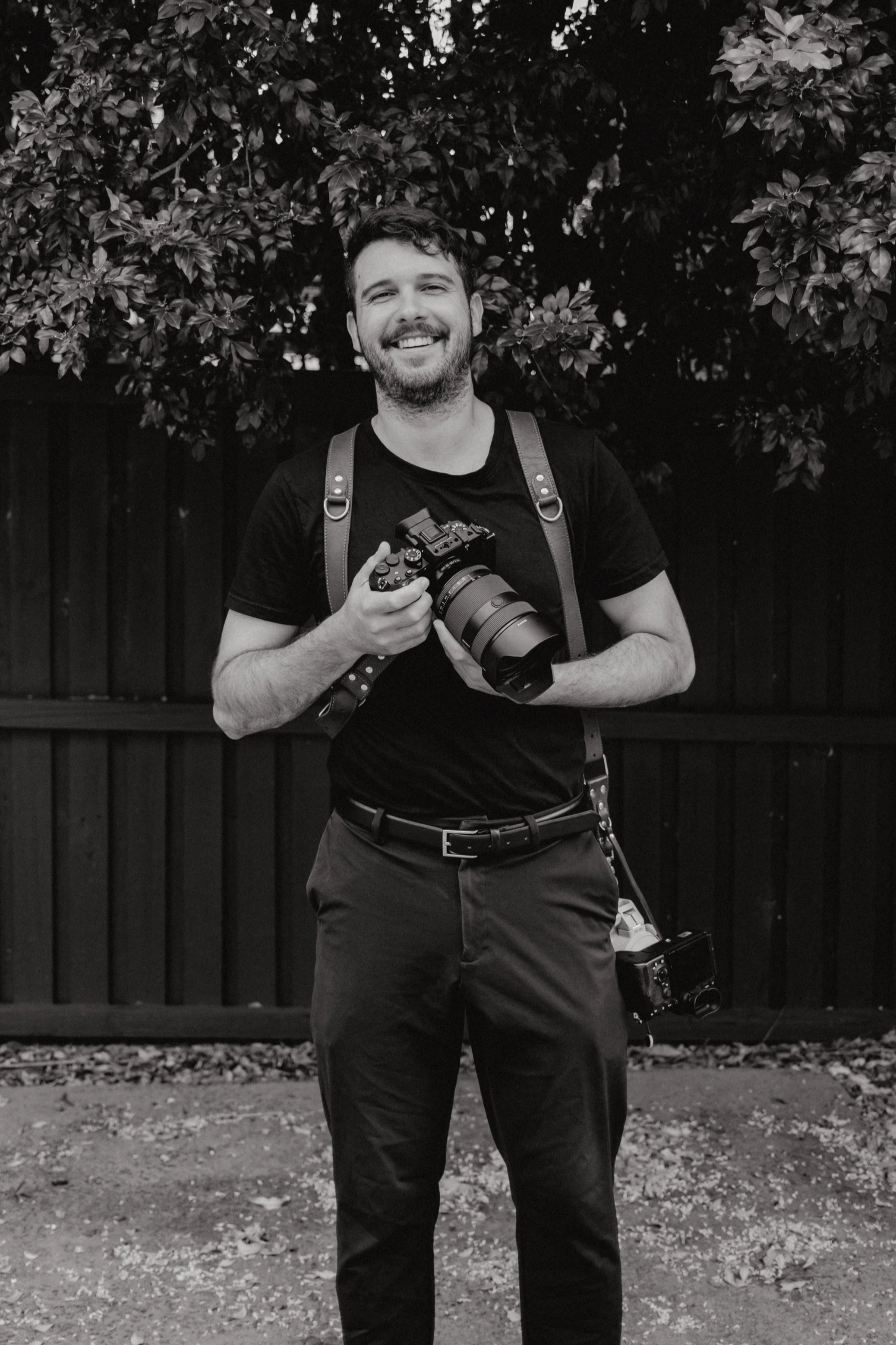A man with a beard smiling, holding a camera, standing outdoors in front of a wooden fence and a large leafy tree, with smaller cameras on the ground near his feet.