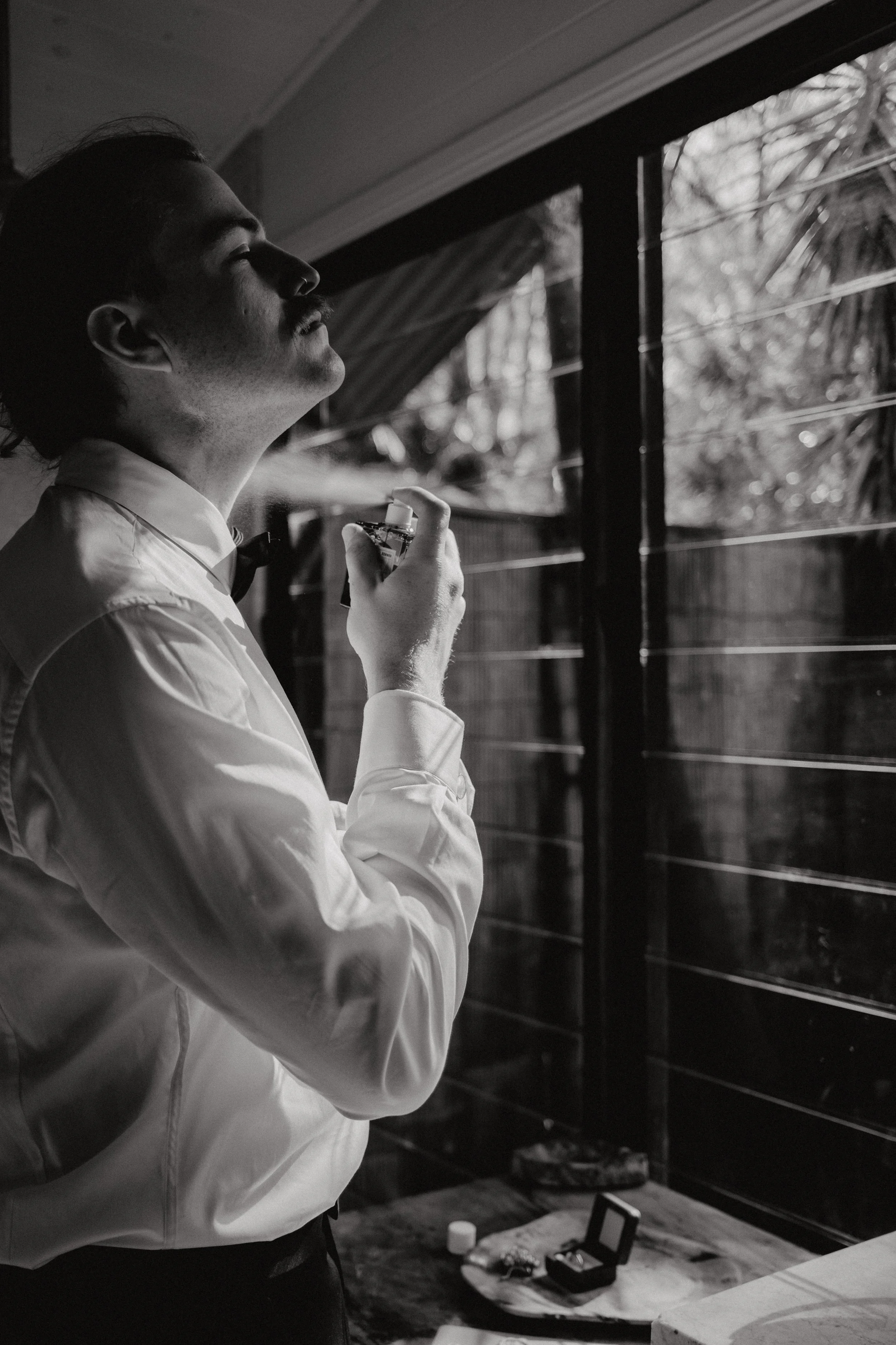 A man wearing a white dress shirt and black bow tie is holding a perfume bottle near his face, standing next to a window with blinds. Sunlight filters through the window, casting shadows on his face and body. There is a table with an open jewelry box