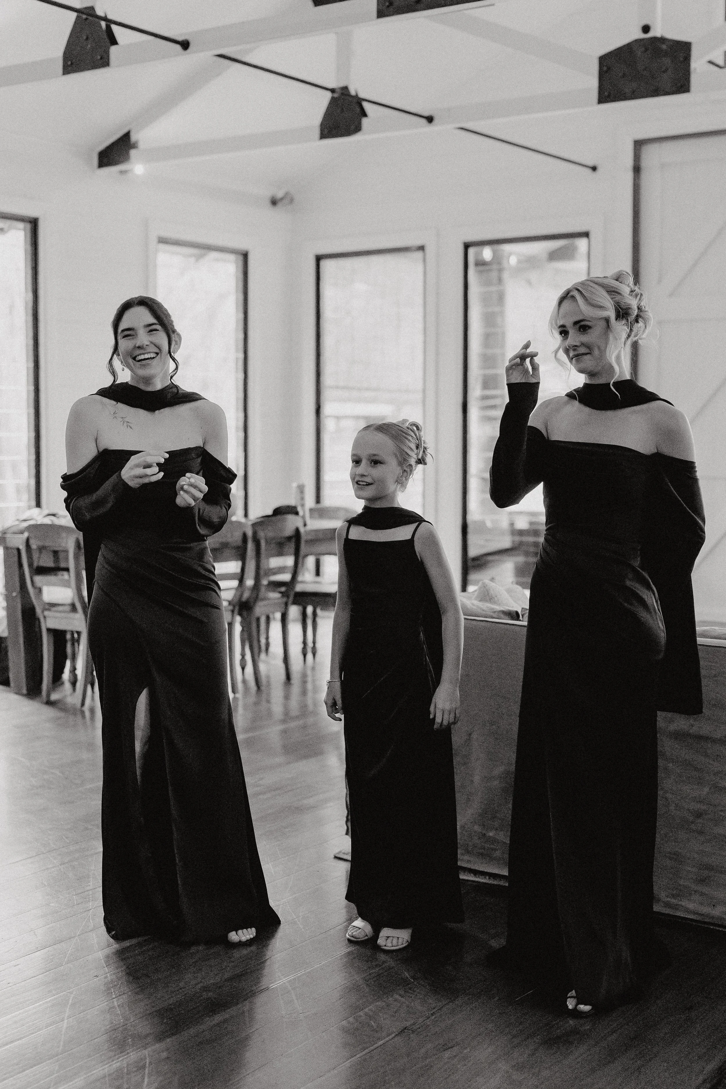 Three women dressed in black formal gowns standing inside a room with large windows. One woman is smiling and talking, another is smiling slightly, and the third has a contemplative expression. There are chairs and tables in the background.