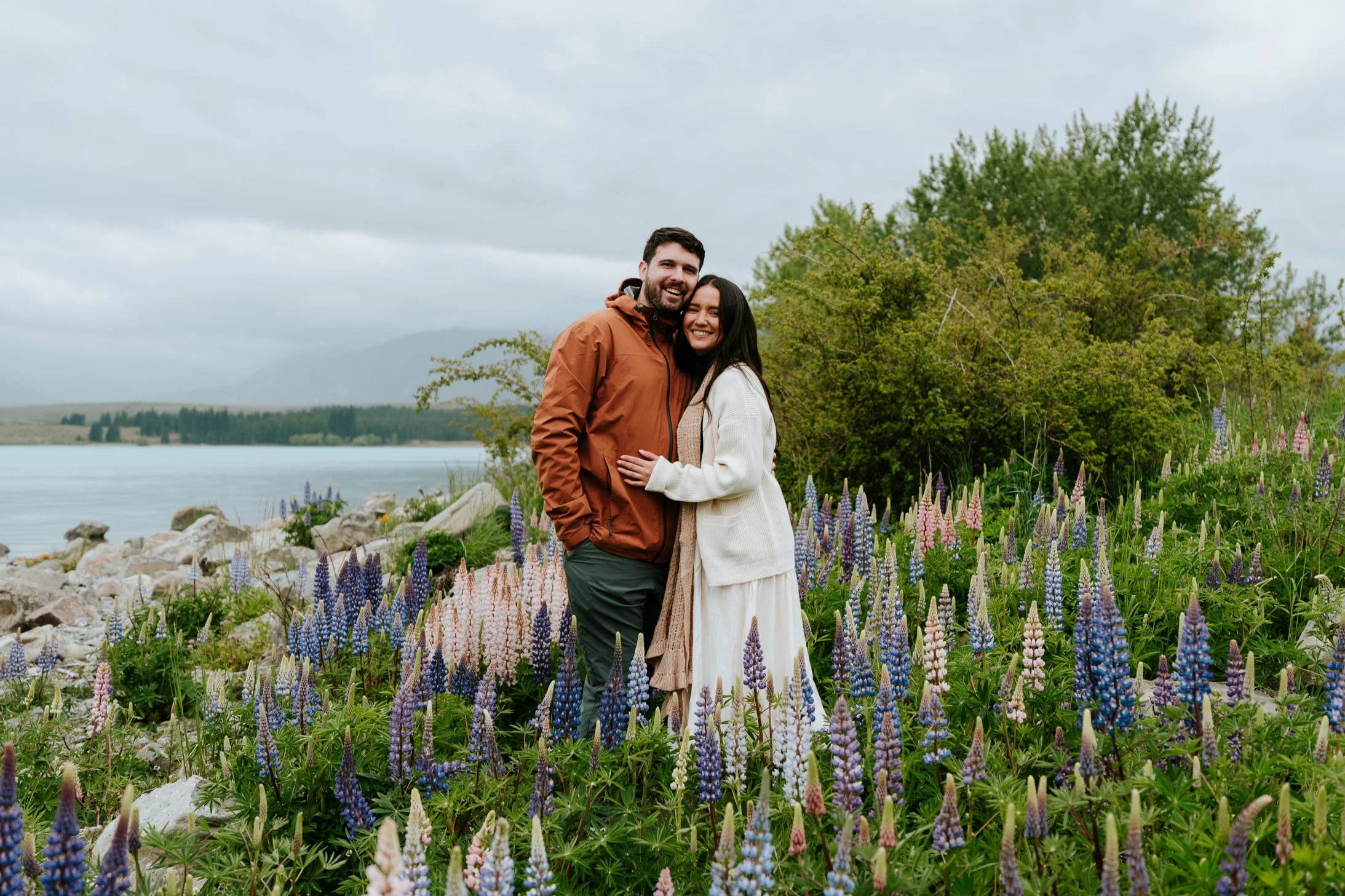 A smiling couple standing amidst vibrant lupine flowers by a lake with mountains and cloudy sky in the background.