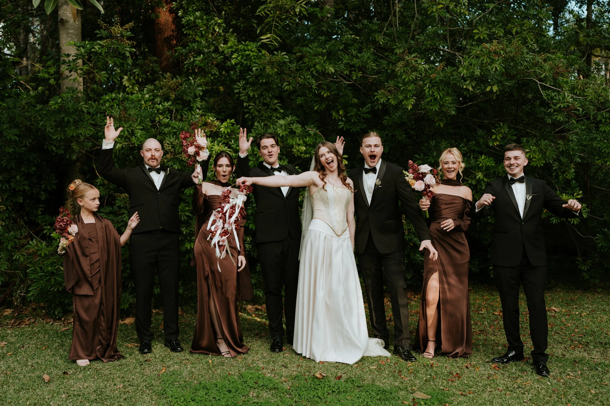 A group of wedding guests in formal attire posing outdoors in front of green foliage, with some holding bouquets and celebrating.
