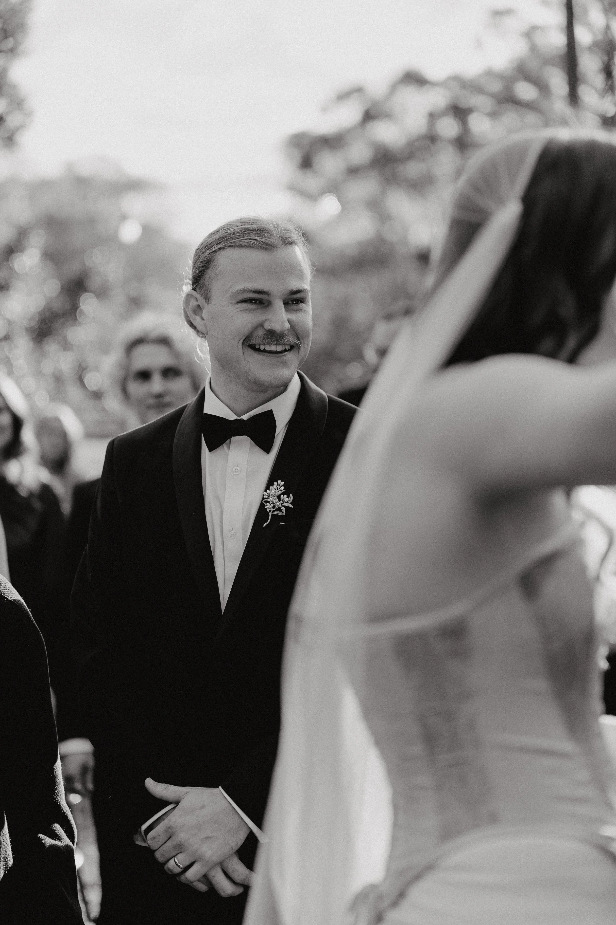 A smiling man in a tuxedo holding hands with a woman in a wedding dress during a wedding ceremony.