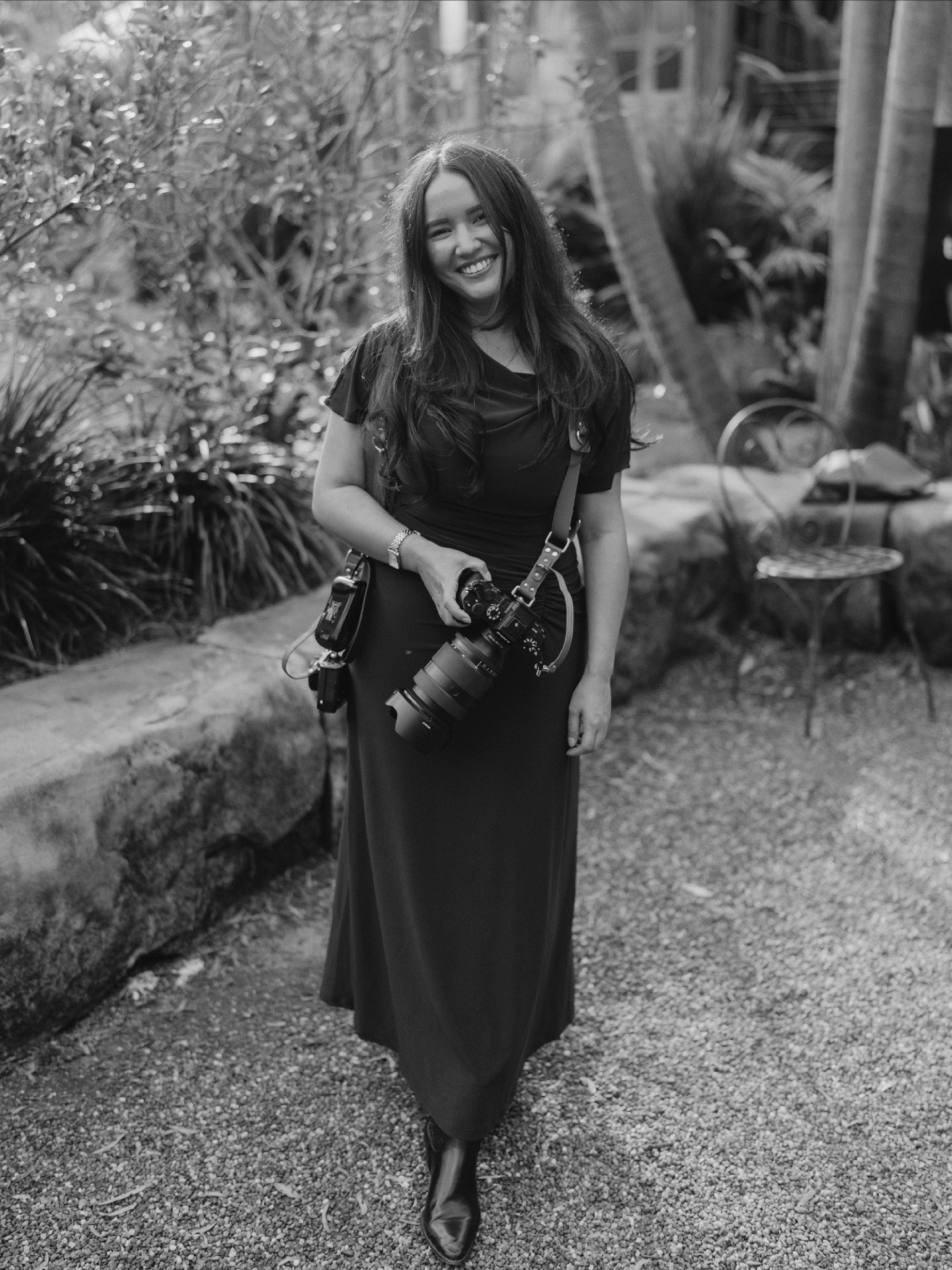 A woman with long hair, wearing a dark dress, smiling and holding a camera, walking outdoors in a garden with trees and plants.