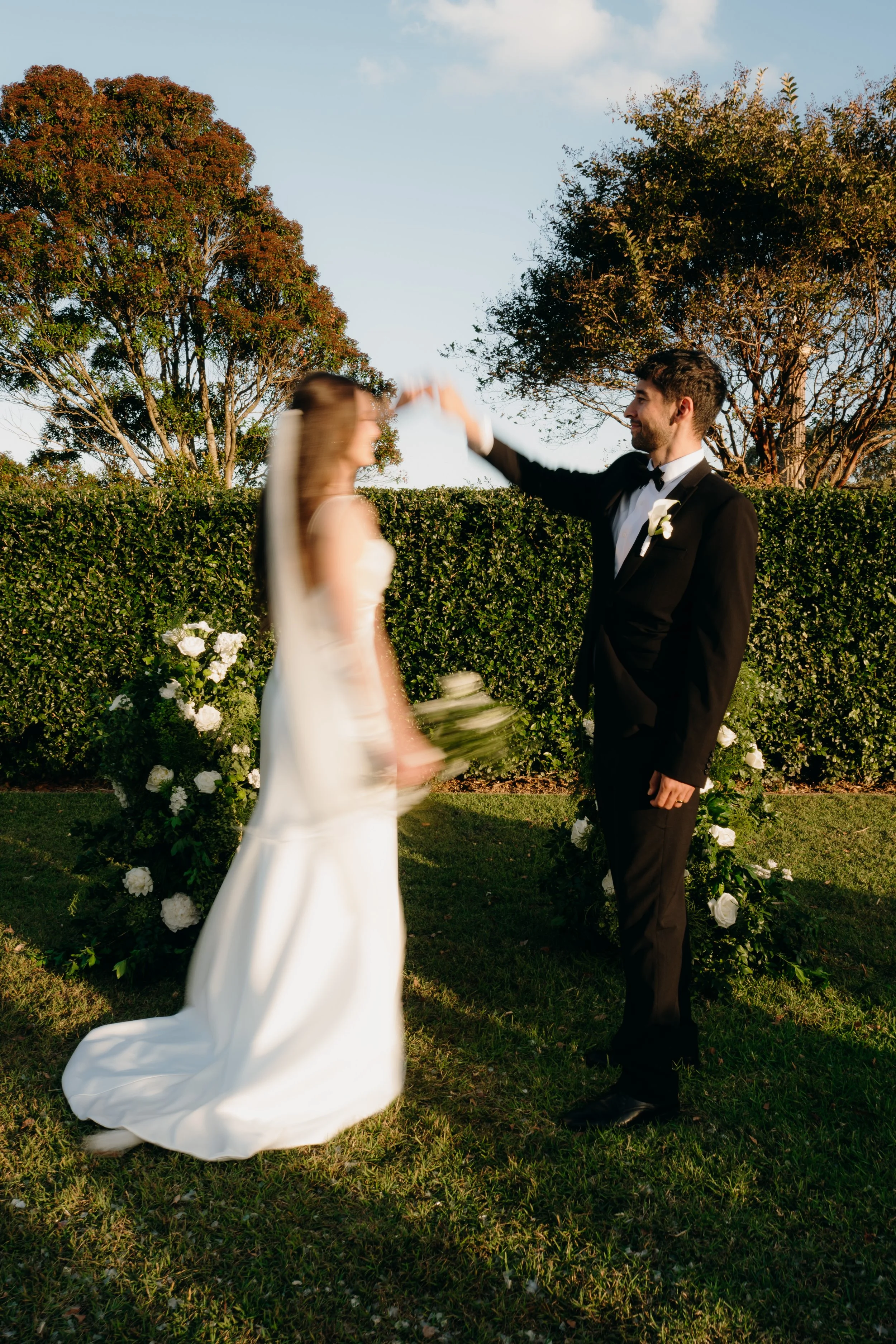 A bride and groom in wedding attire facing each other outdoors, holding hands, with greenery and trees in the background, during sunset.