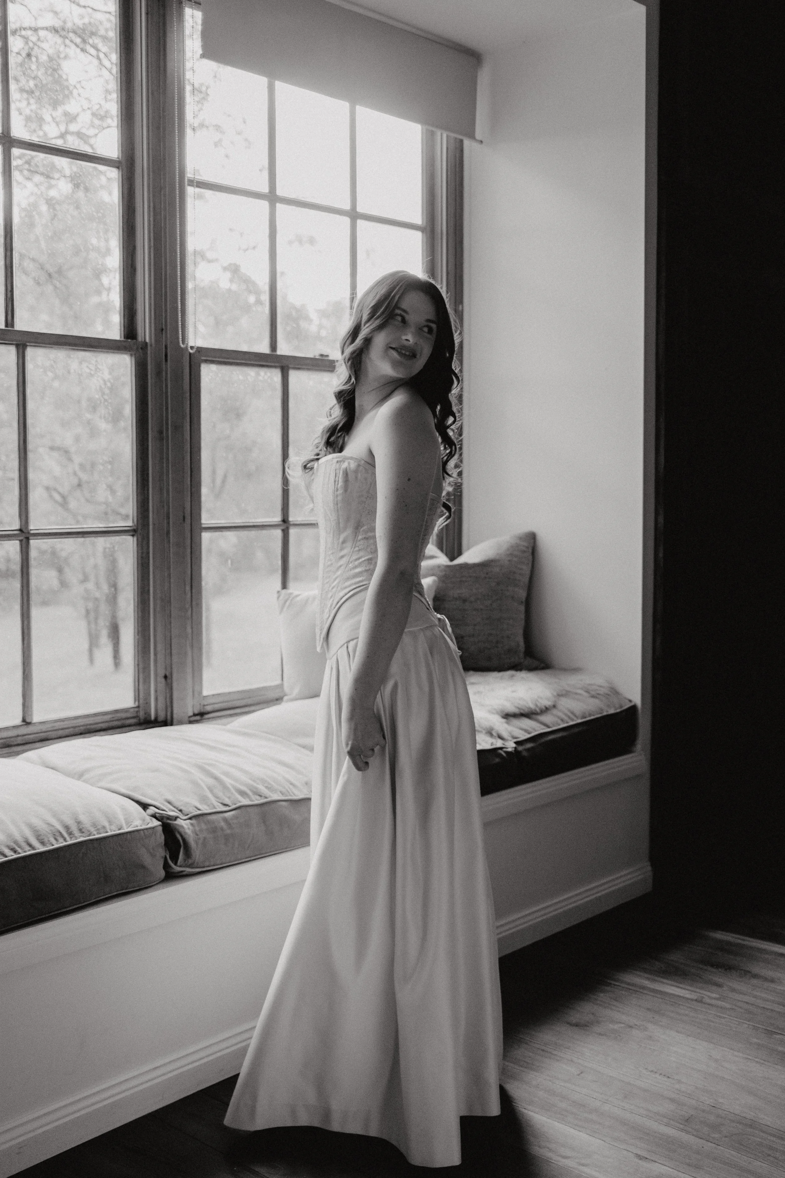 A woman in a strapless gown stands next to a bed with cushions, by a large window with multiple panes, in a room with carpeted floors, during daytime.