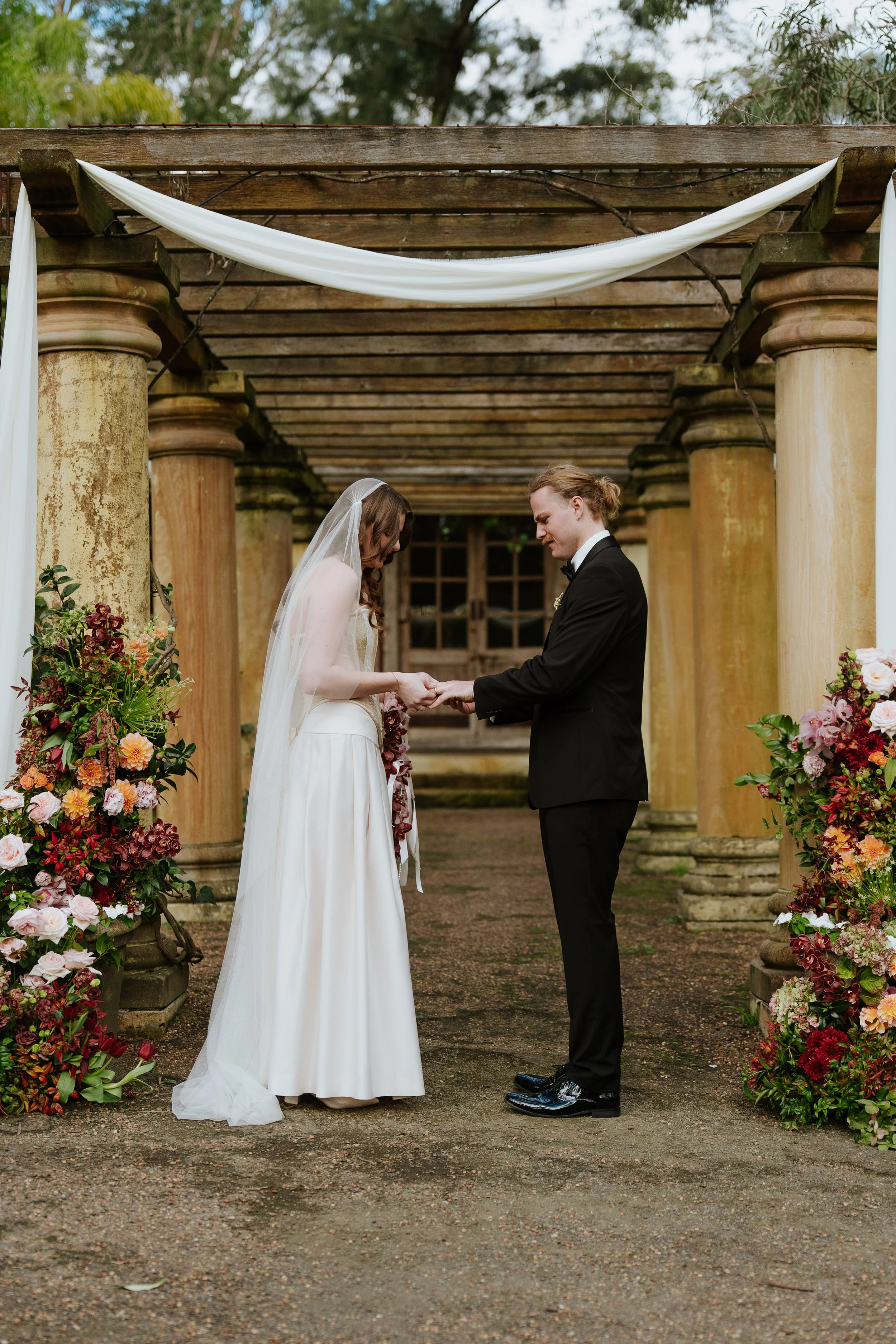 A bride and groom holding hands during a wedding ceremony outdoors, under a wooden pergola decorated with white fabric and floral arrangements, with green trees in the background.