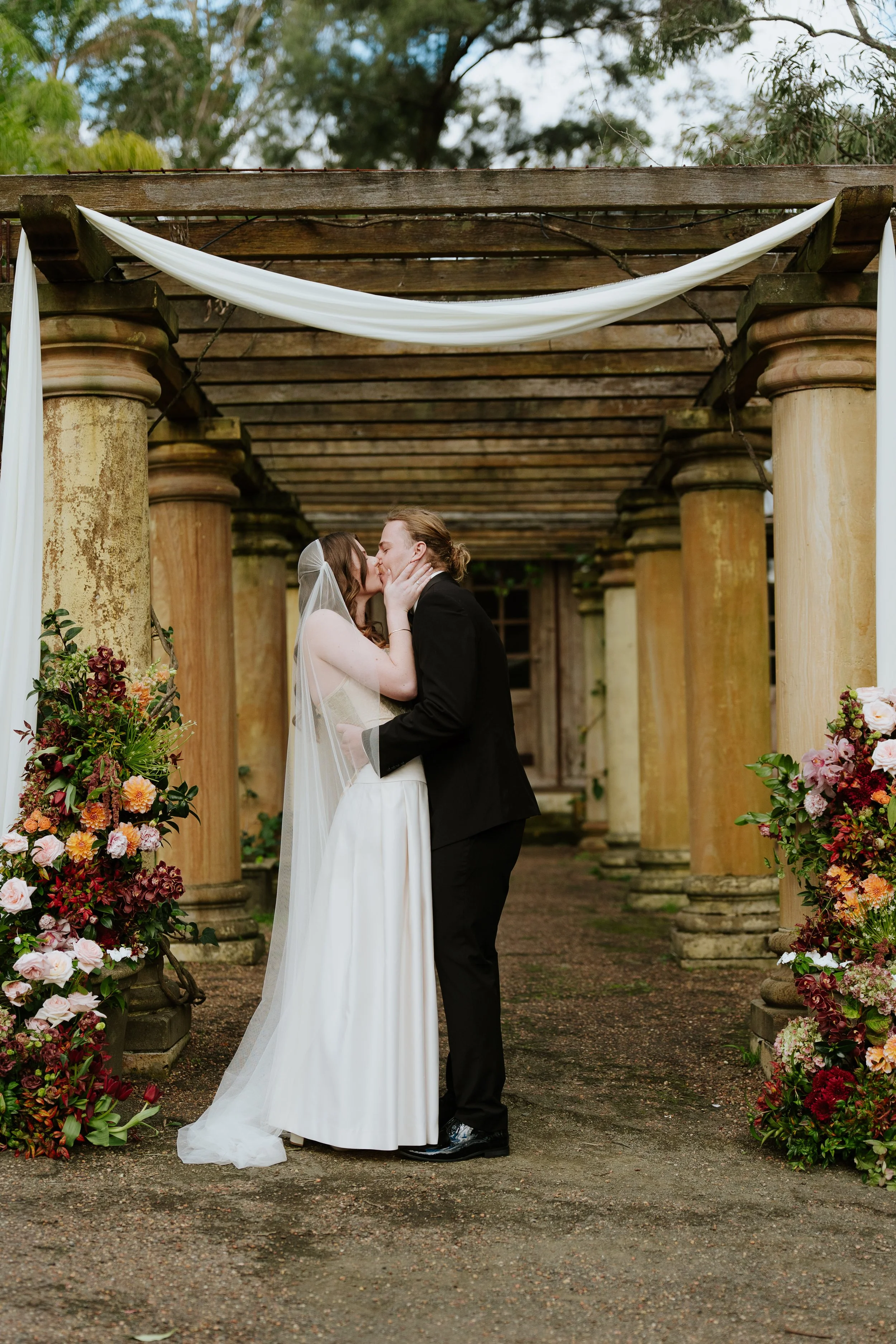 A bride and groom kissing under a wooden pergola decorated with draped white fabric and surrounded by colorful flower arrangements.
