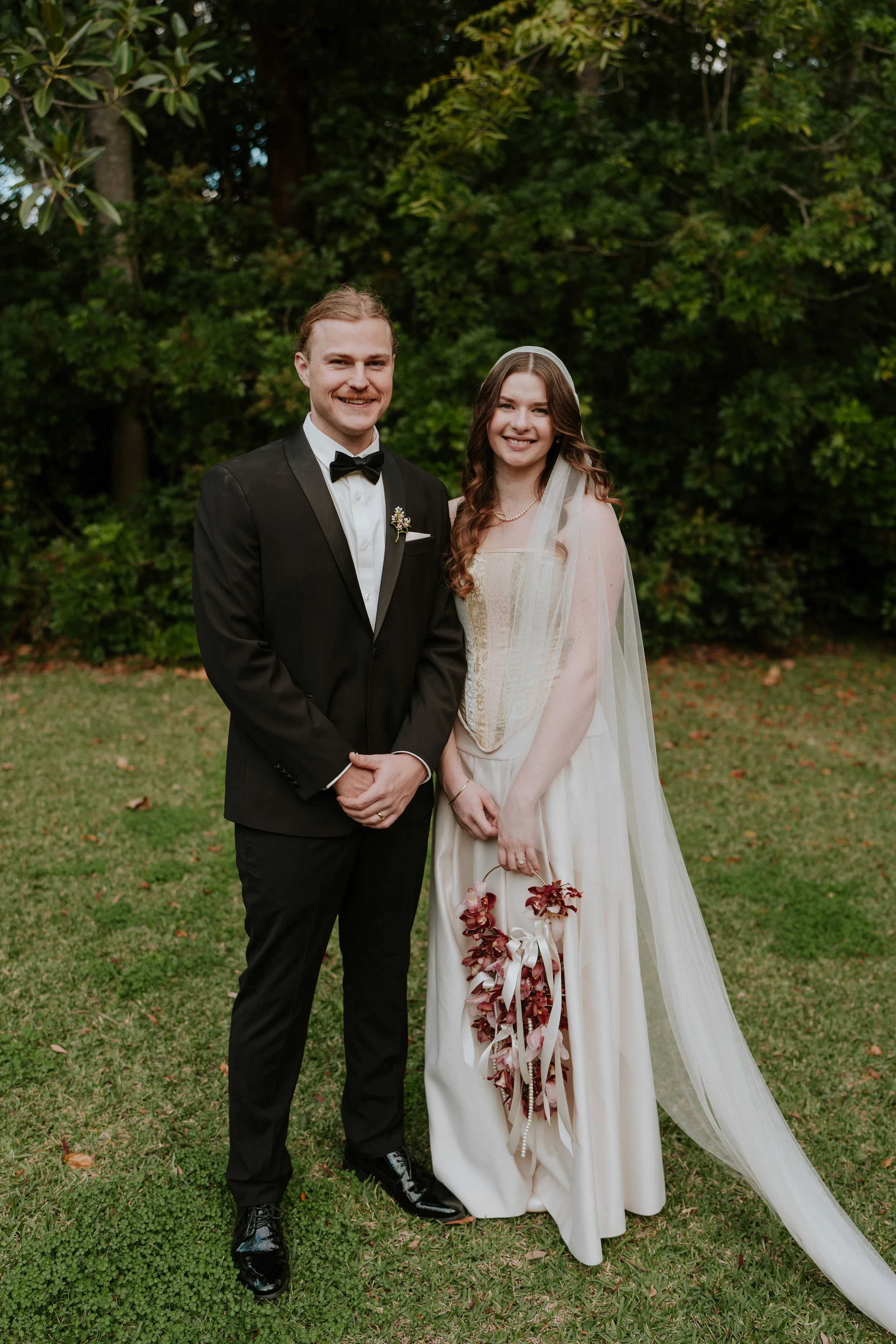 A smiling couple dressed in wedding attire standing on a grassy area outdoors with lush green trees in the background. The groom is wearing a black tuxedo with a bow tie, and the bride is wearing a cream-colored wedding dress with a sheer veil and holding a bouquet of flowers.