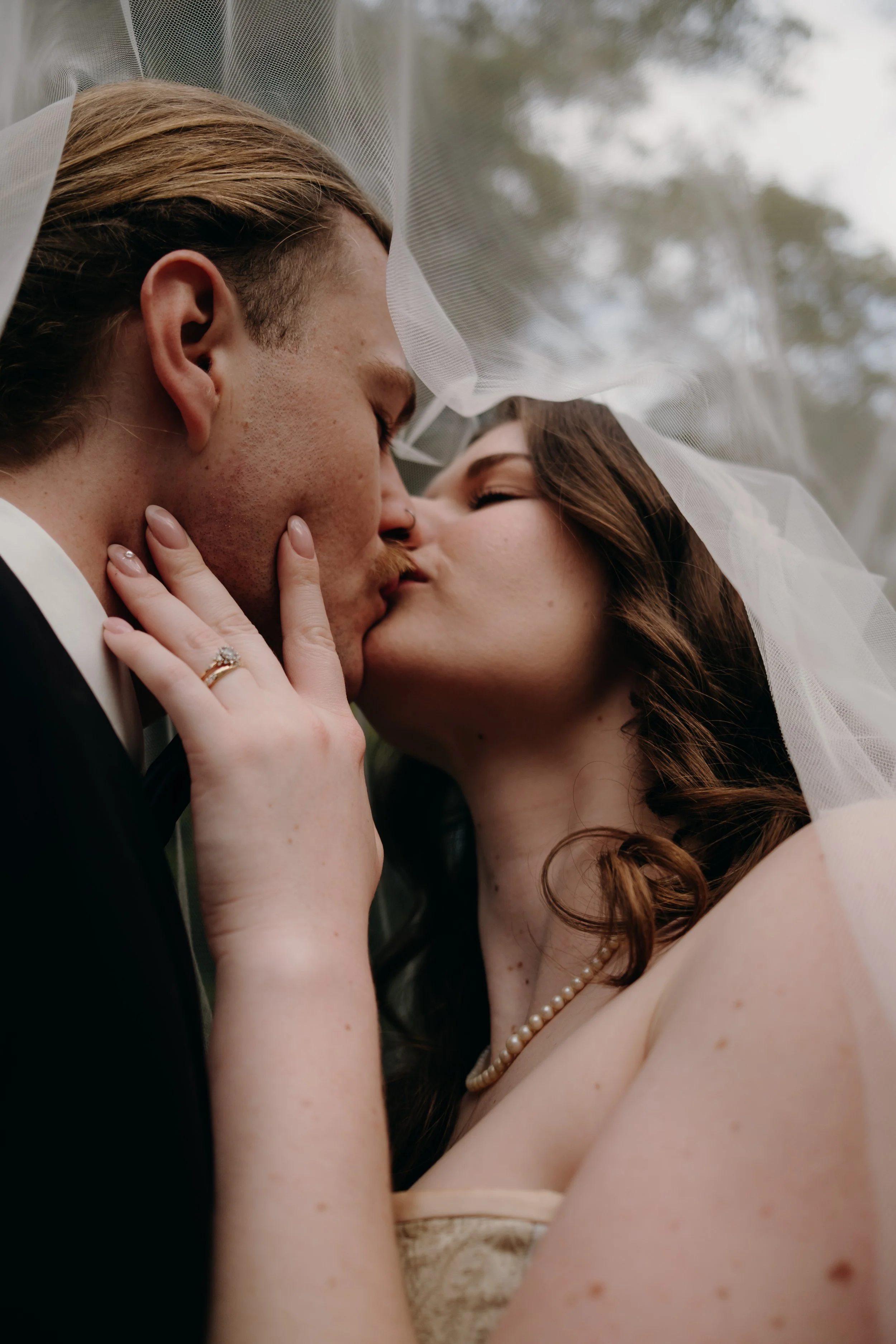 A close-up photo of a bride and groom kissing with a sheer veil over their faces. The bride has red hair, is wearing a pearl necklace and ring, and has her hand on the groom’s face. The groom has blond hair, a side part, and is dressed in a tuxedo wi