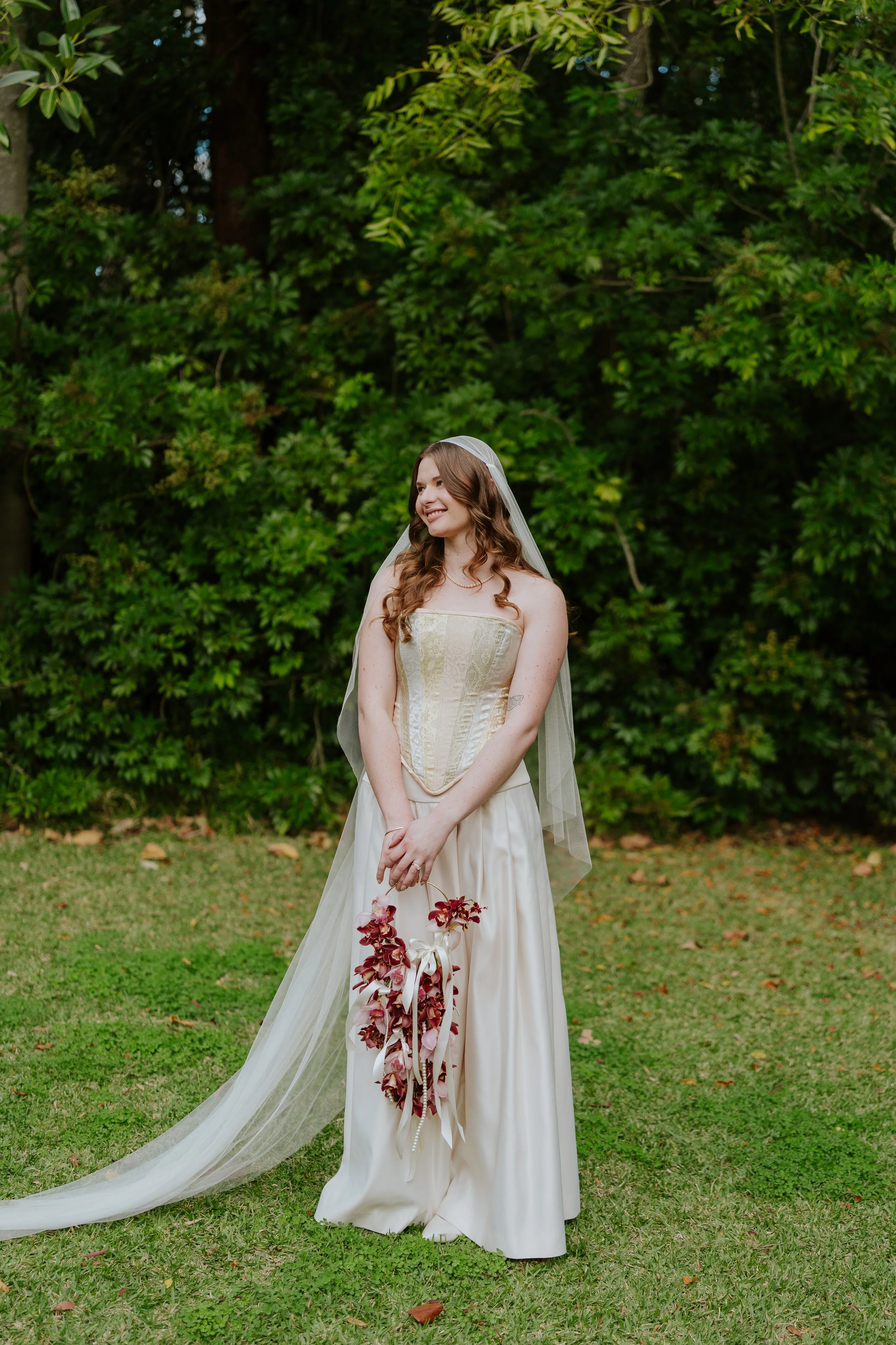 A young woman in a white wedding dress and veil stands outdoors on grass, holding a bouquet of pink and white flowers, smiling and looking to her left, with lush green trees in the background.