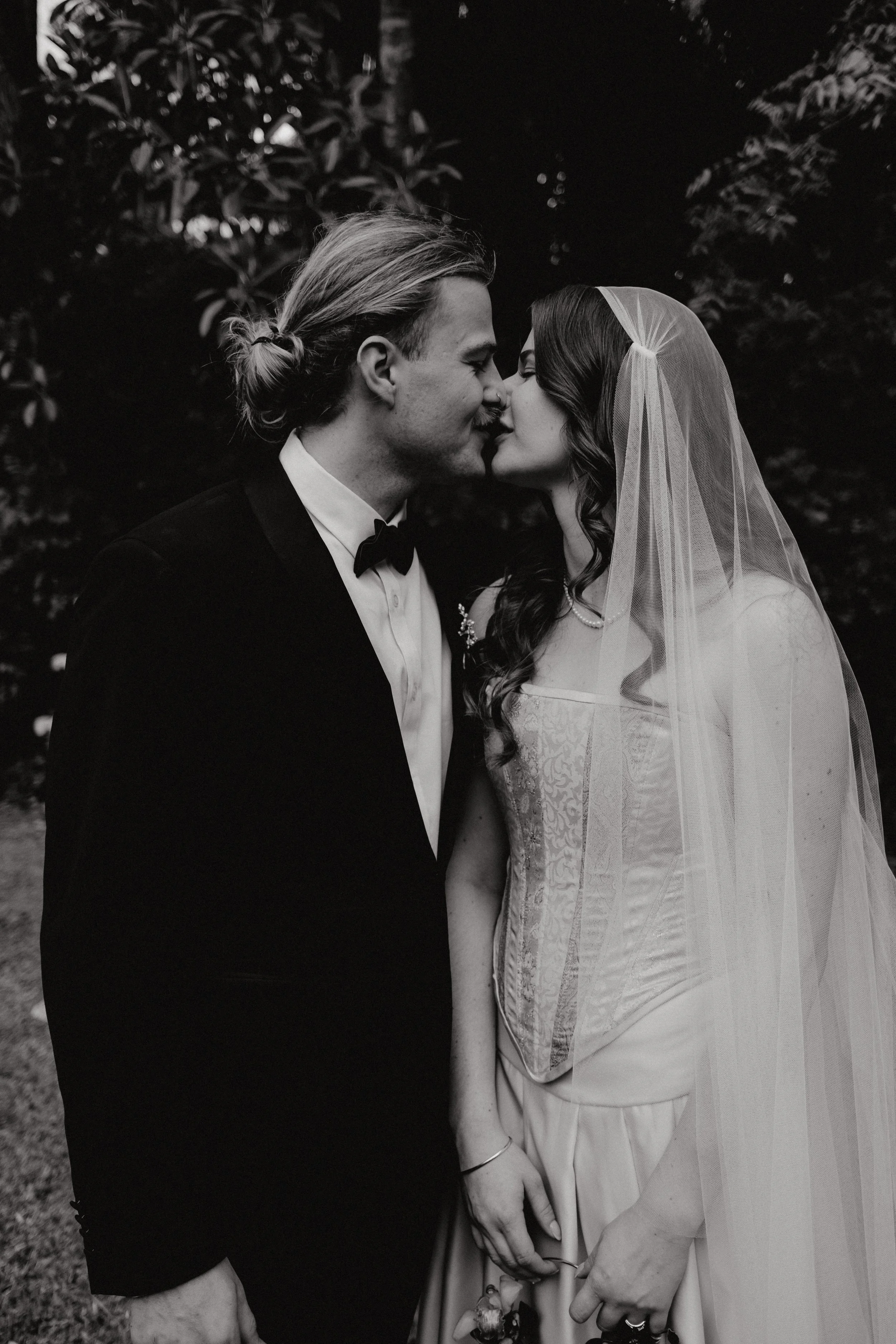 A black and white photograph of a bride and groom standing close together, with their foreheads and noses touching. The groom is wearing a tuxedo with a bow tie, and the bride is wearing a wedding dress with a lace bodice and a veil. They are holding hands, and the background consists of trees and foliage.