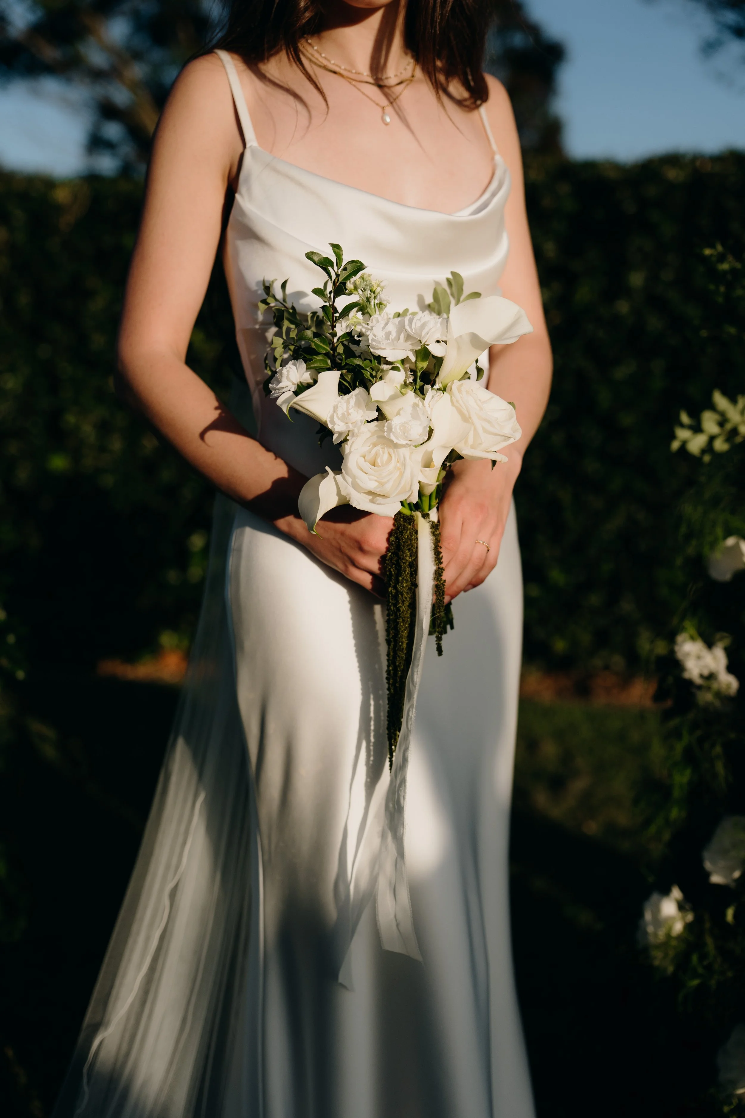 A woman in a white dress holding a bouquet of white flowers and greenery outdoors.