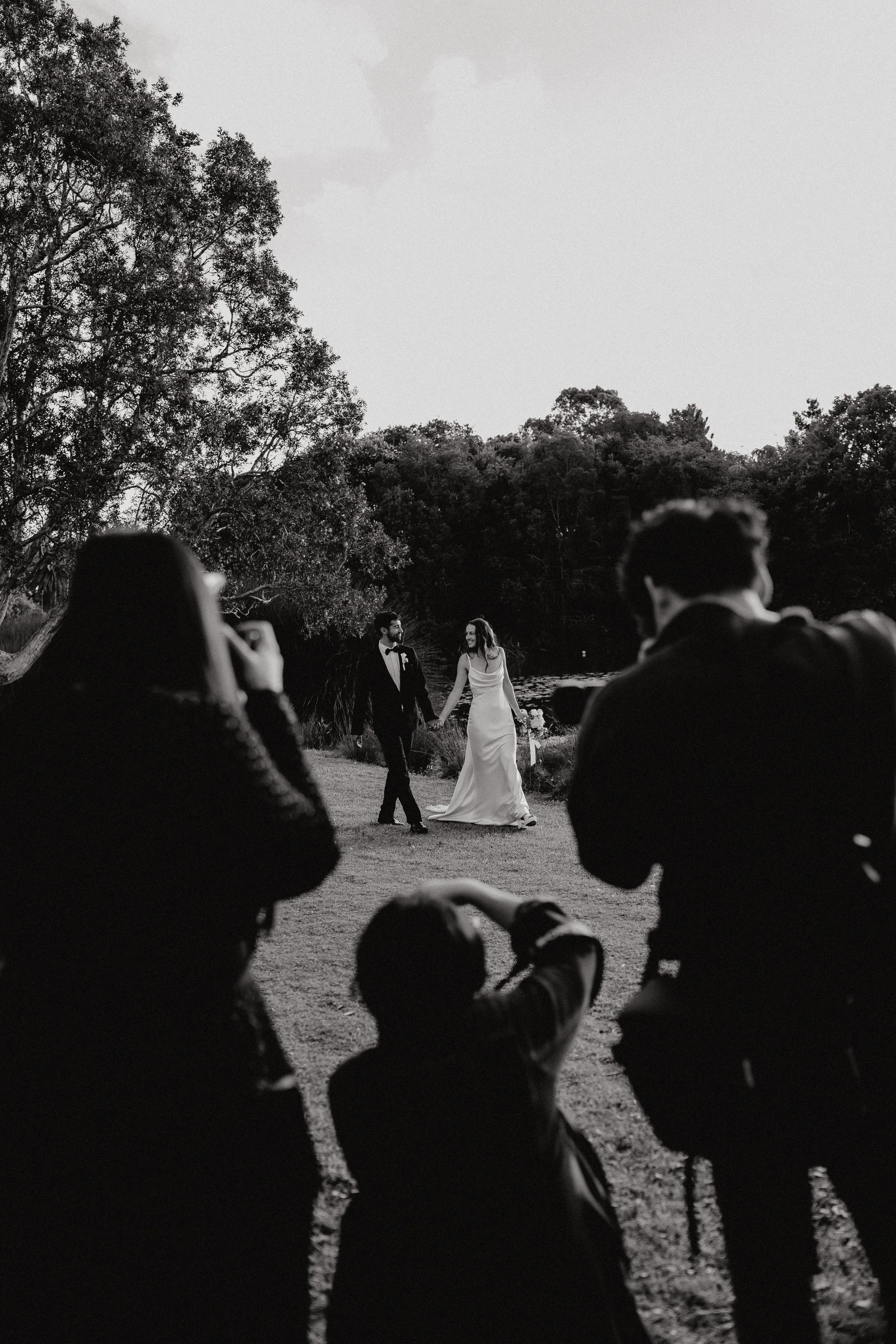 Black and white photo of a bride and groom holding hands, walking outdoors near a river, with guests sitting in the foreground taking pictures. The scene is surrounded by trees and appears to be part of a wedding celebration.