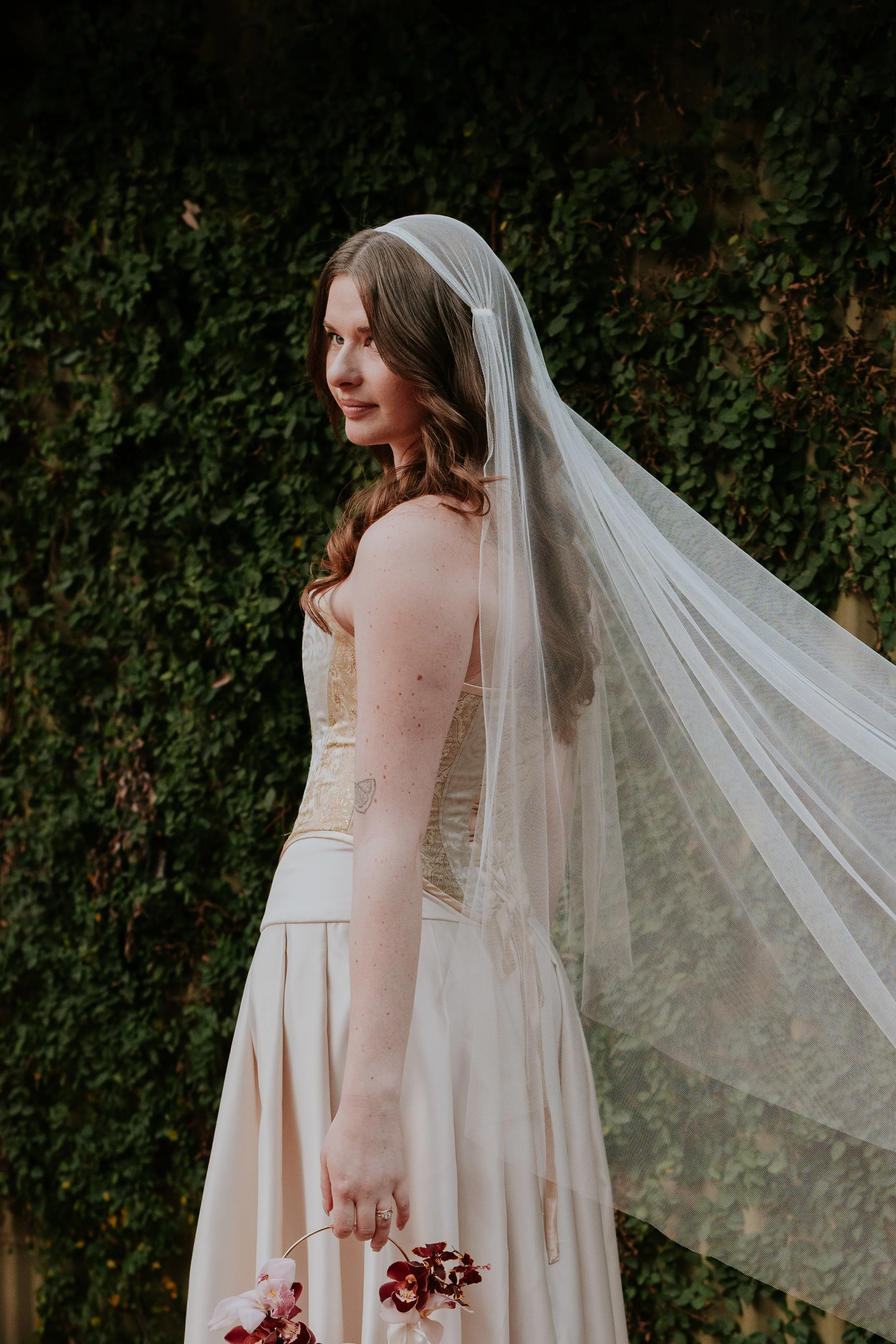 A woman with brown hair in loose curls wearing a wedding dress and veil standing outdoors against a green foliage background, holding a small floral bouquet.