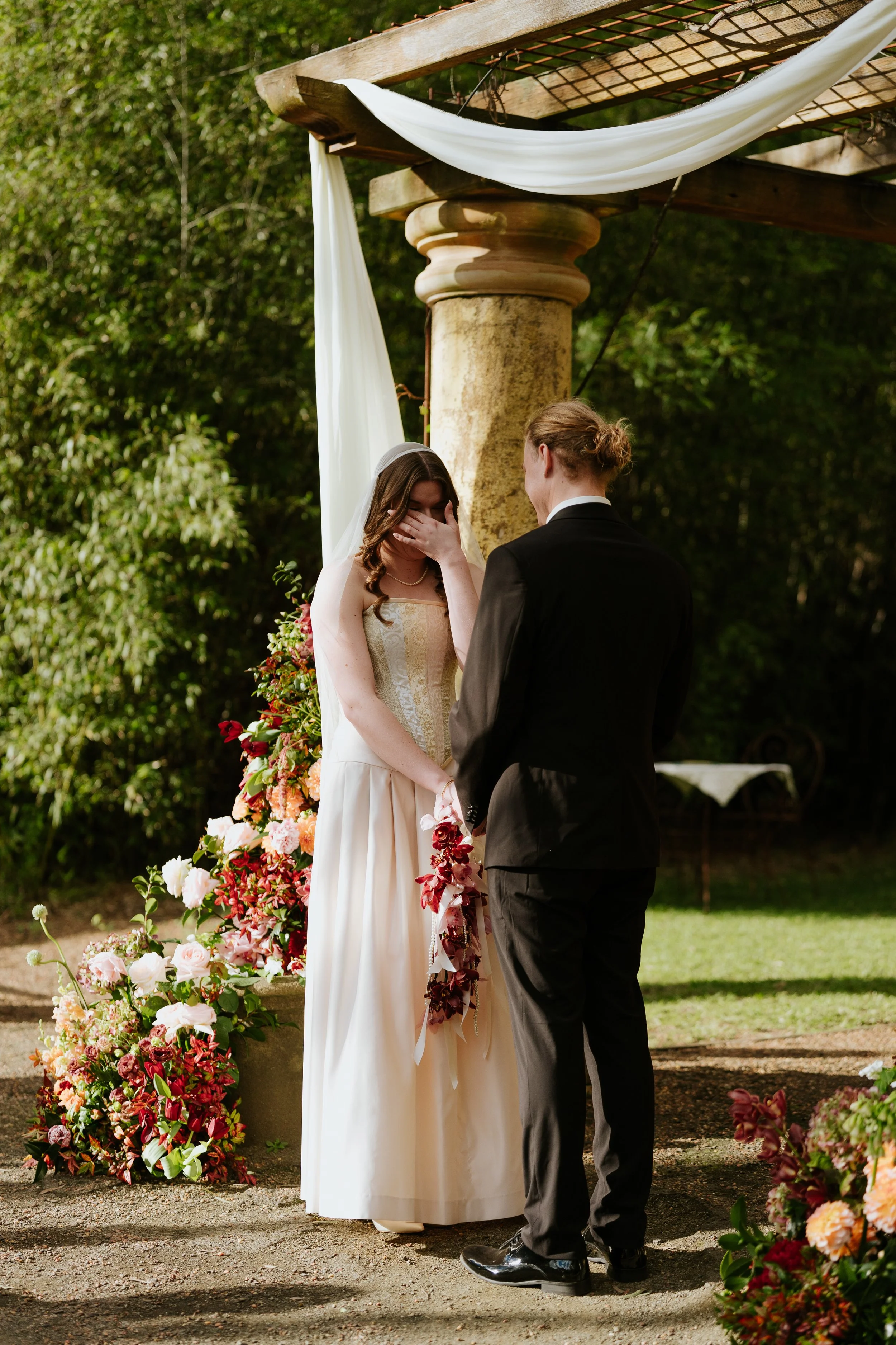 A bride and groom are exchanging vows during an outdoor wedding ceremony. The bride is covering her face with her hand, and the groom is facing her. They are standing in front of a floral arrangement with pink, red, and white flowers, and a white fab