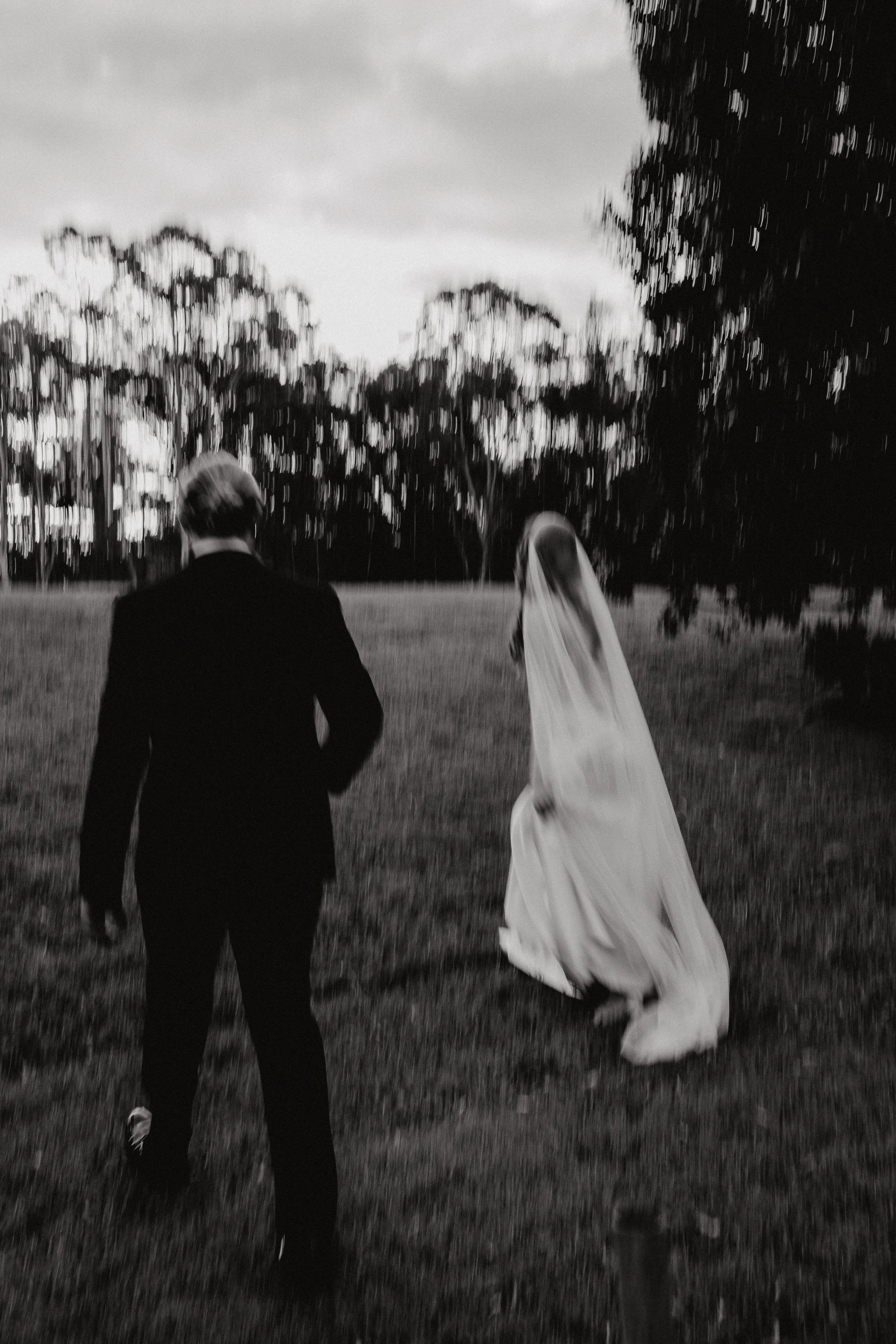 A black-and-white photo of a couple walking hand in hand in a park or outdoor area, with trees in the background. The woman is wearing a long dress and a headscarf, and the man is dressed in a suit.