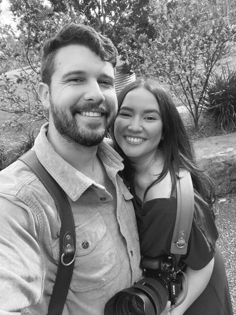A happy couple taking a selfie outdoors, with greenery in the background, both smiling at the camera. The man has a beard and is wearing a light shirt with a camera hanging from his neck, and the woman has long dark hair and is also smiling.