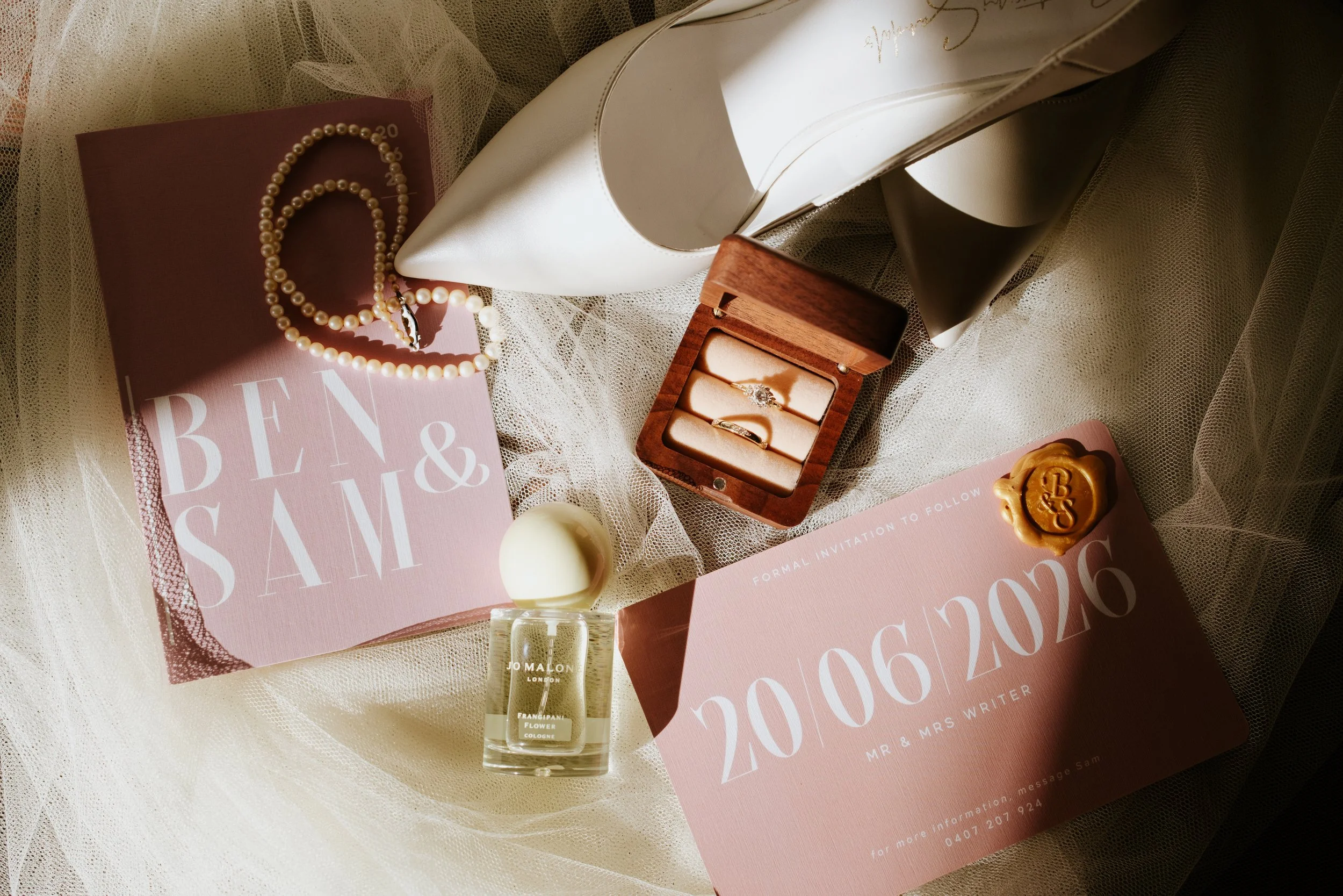 Various wedding accessories including a pearl necklace, a pair of pearl earrings, a box with rings, a bottle of Jo Malone perfume, candles, and wedding invitations on a lace fabric background.