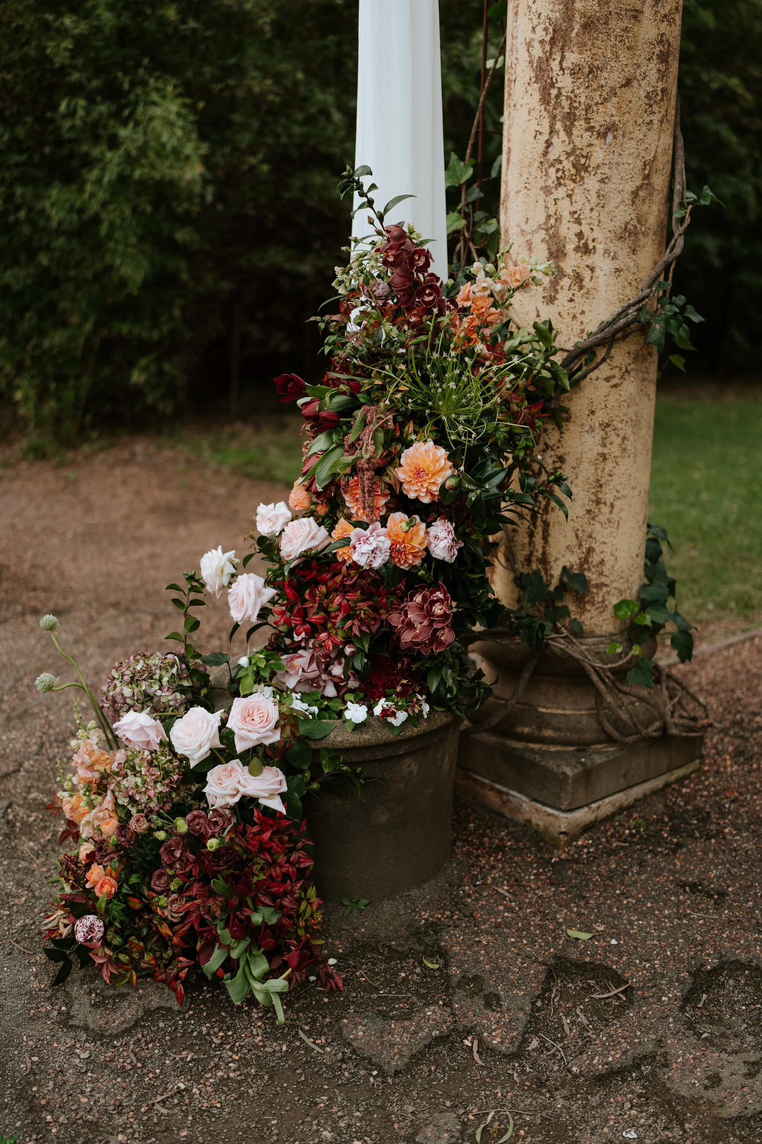 Flower arrangement with pink, peach, white, and dark red flowers placed in large planters at the base of a rustic stone and wood pillar outdoors.