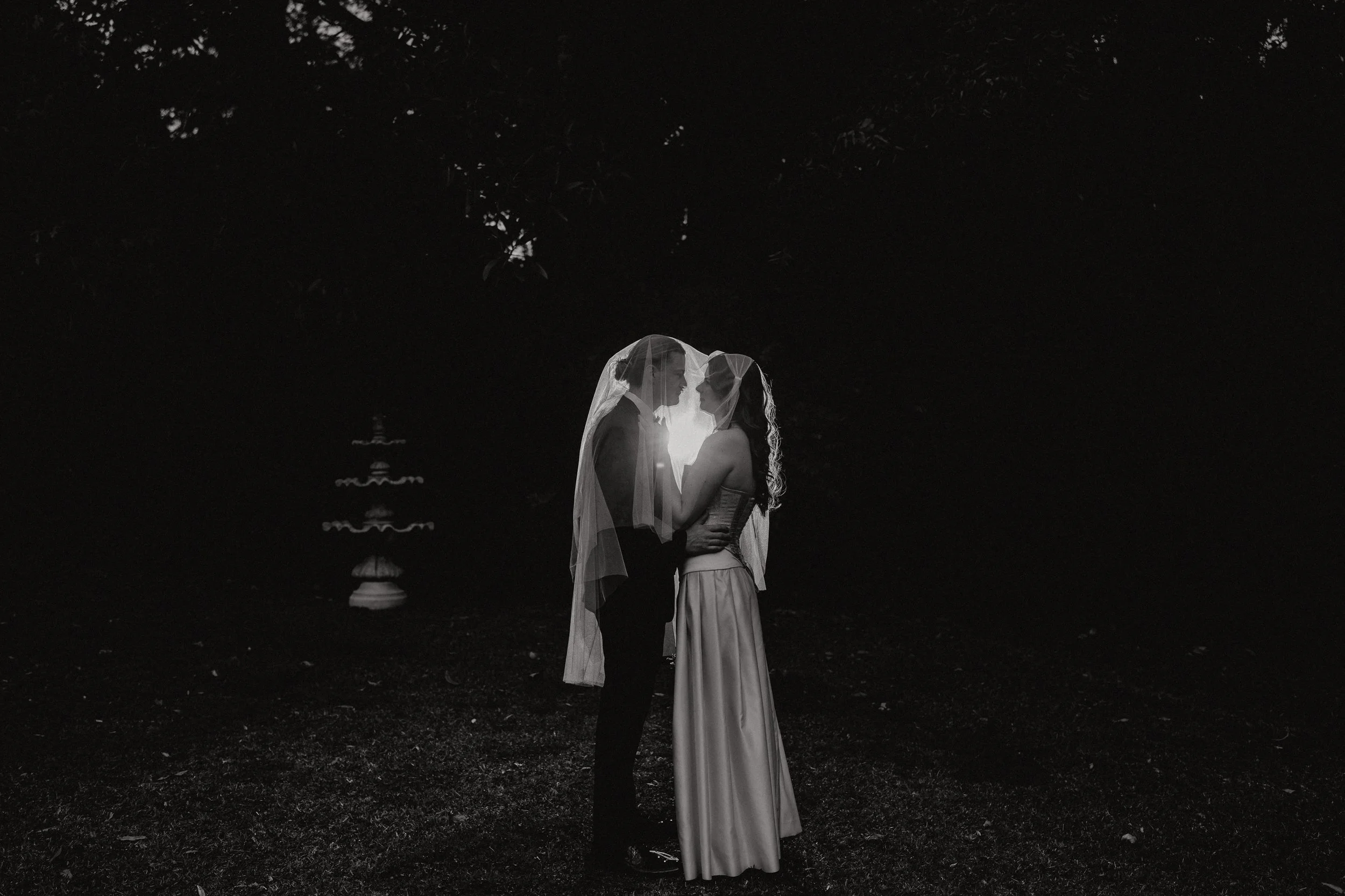A black-and-white photo of a couple dressed in wedding attire, standing close and facing each other under a dark, tree-filled background.
