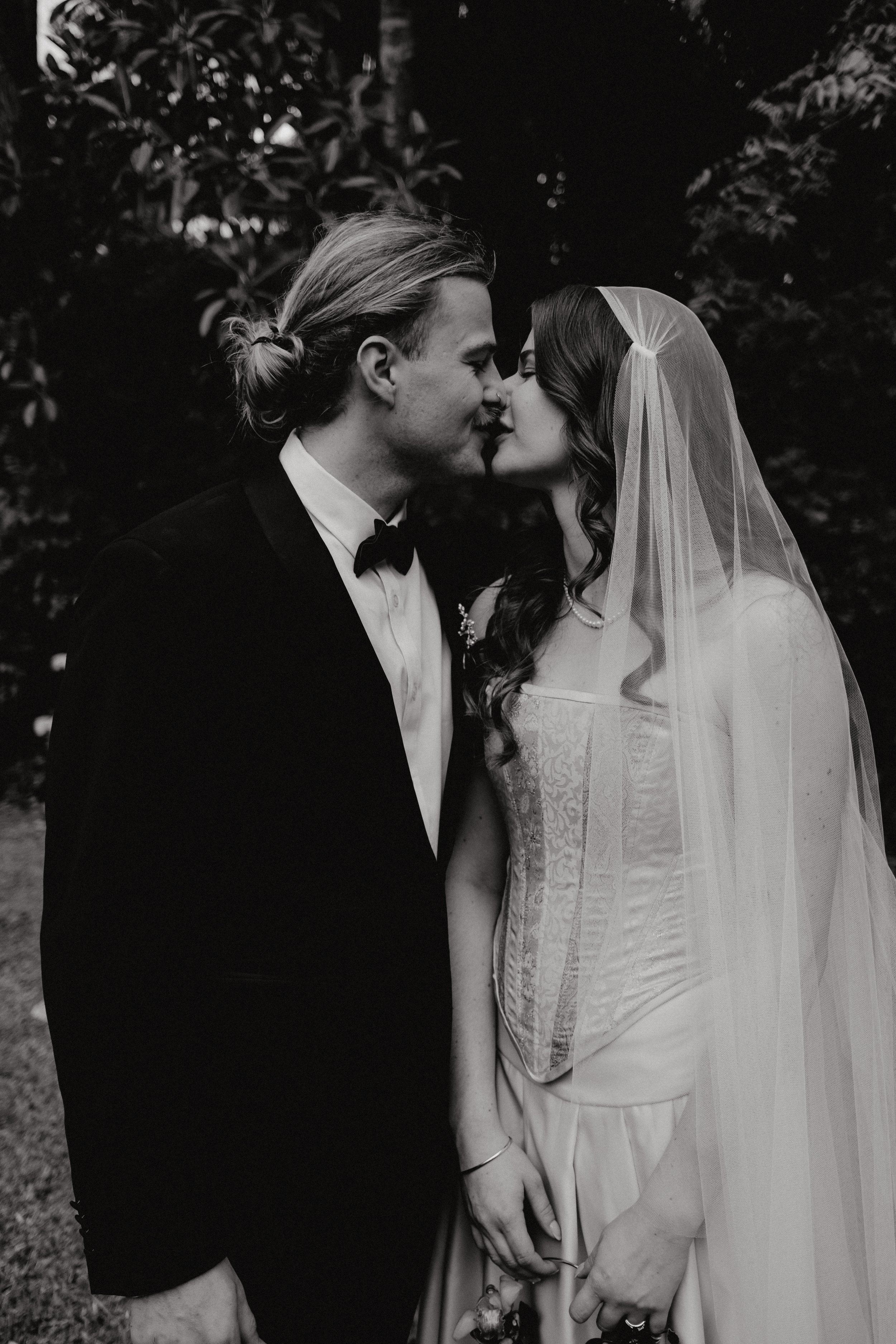 A black and white photograph of a newlywed couple standing close together, gently touching noses, with the bride wearing a lace wedding gown and a veil, and the groom in a tuxedo with a bow tie, holding a bouquet in her hand.