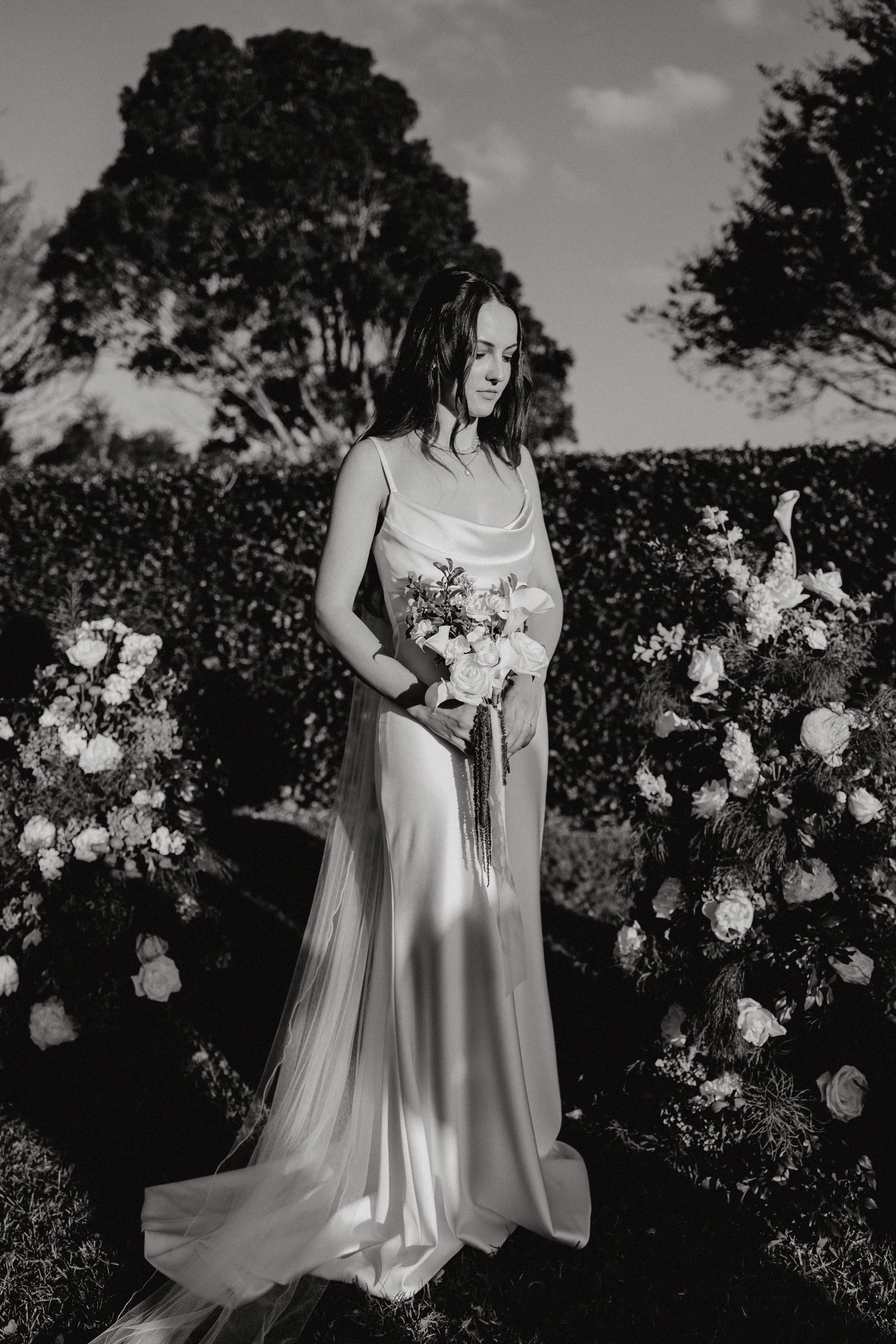 Black and white photo of a young woman in a satin dress holding a bouquet of flowers, standing outdoors in front of a floral arrangement and trees.