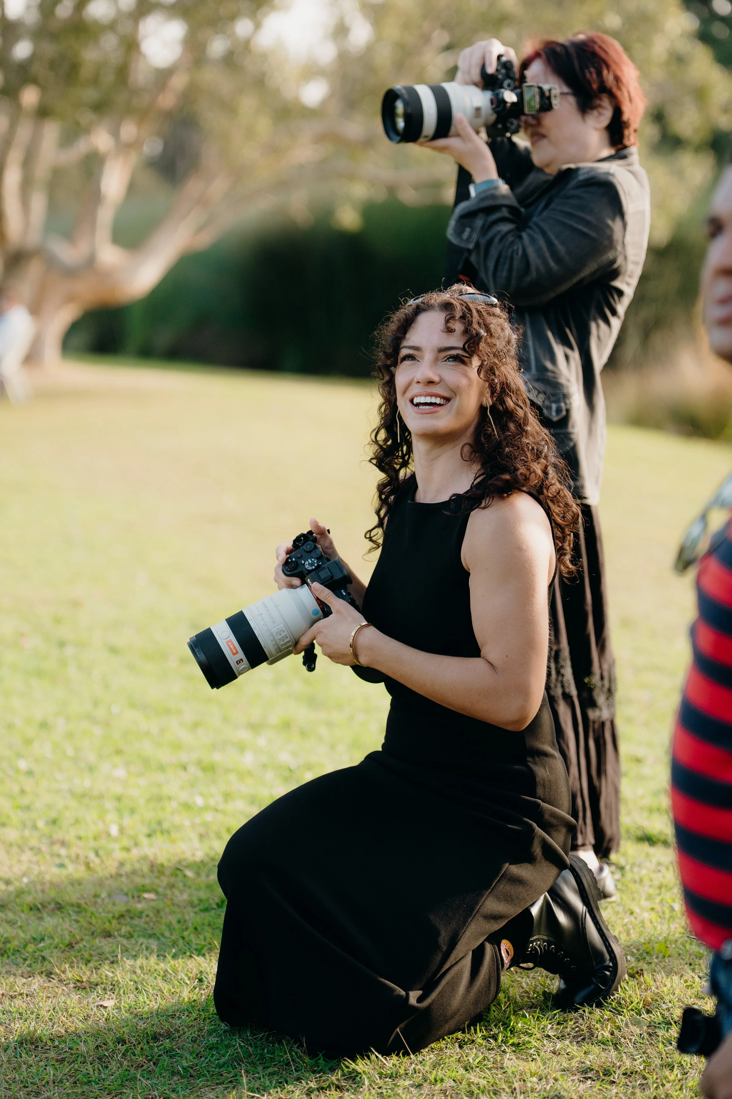 A woman with curly hair and earrings sitting on the grass, holding a camera with a large telephoto lens, smiling and looking up. Behind her, a woman with short red hair and a black jacket is taking a photo with a camera, standing on one knee. Other p