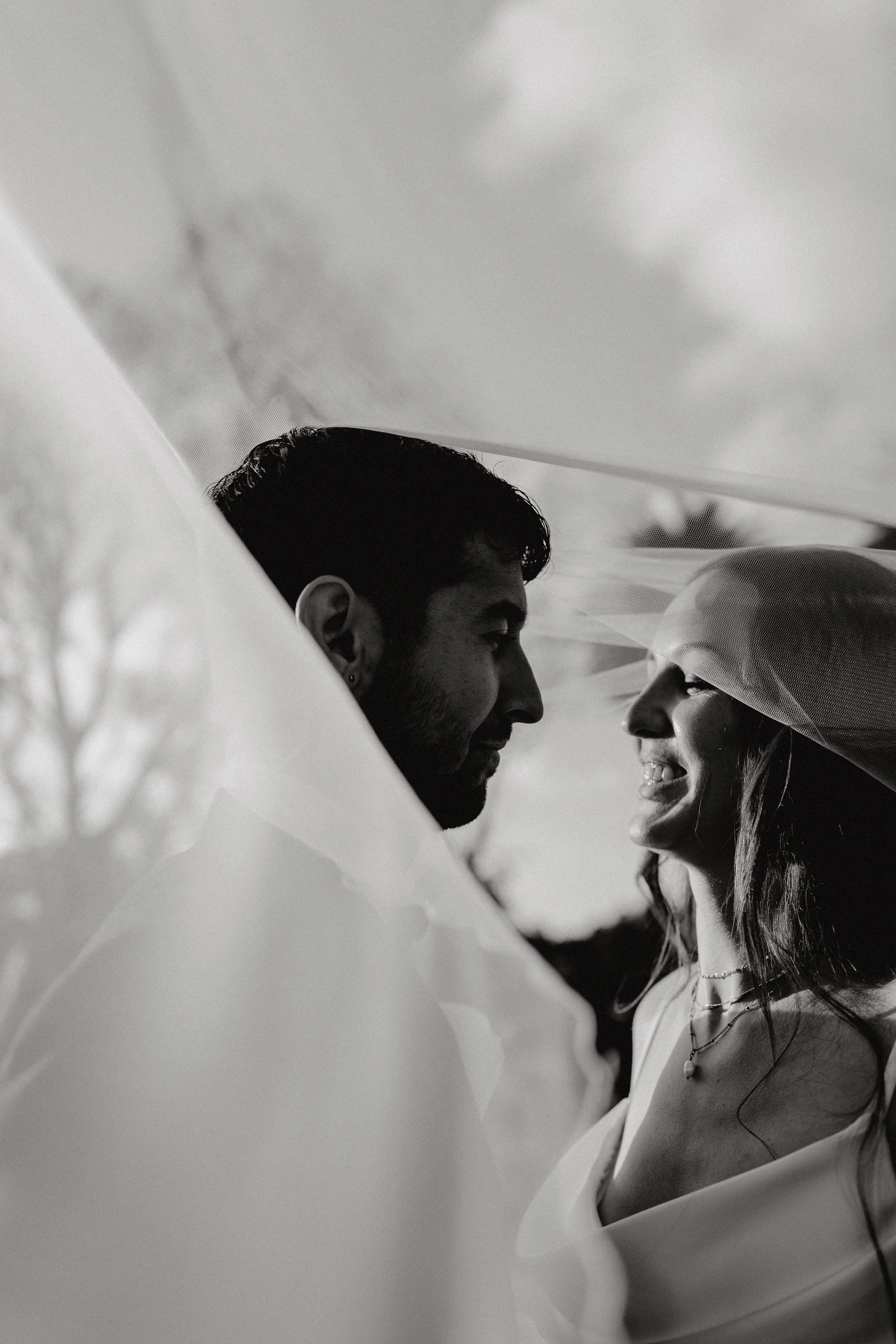 A black and white photo of a man and woman smiling and looking at each other, with the woman wearing a veil, suggesting a wedding or romantic moment.
