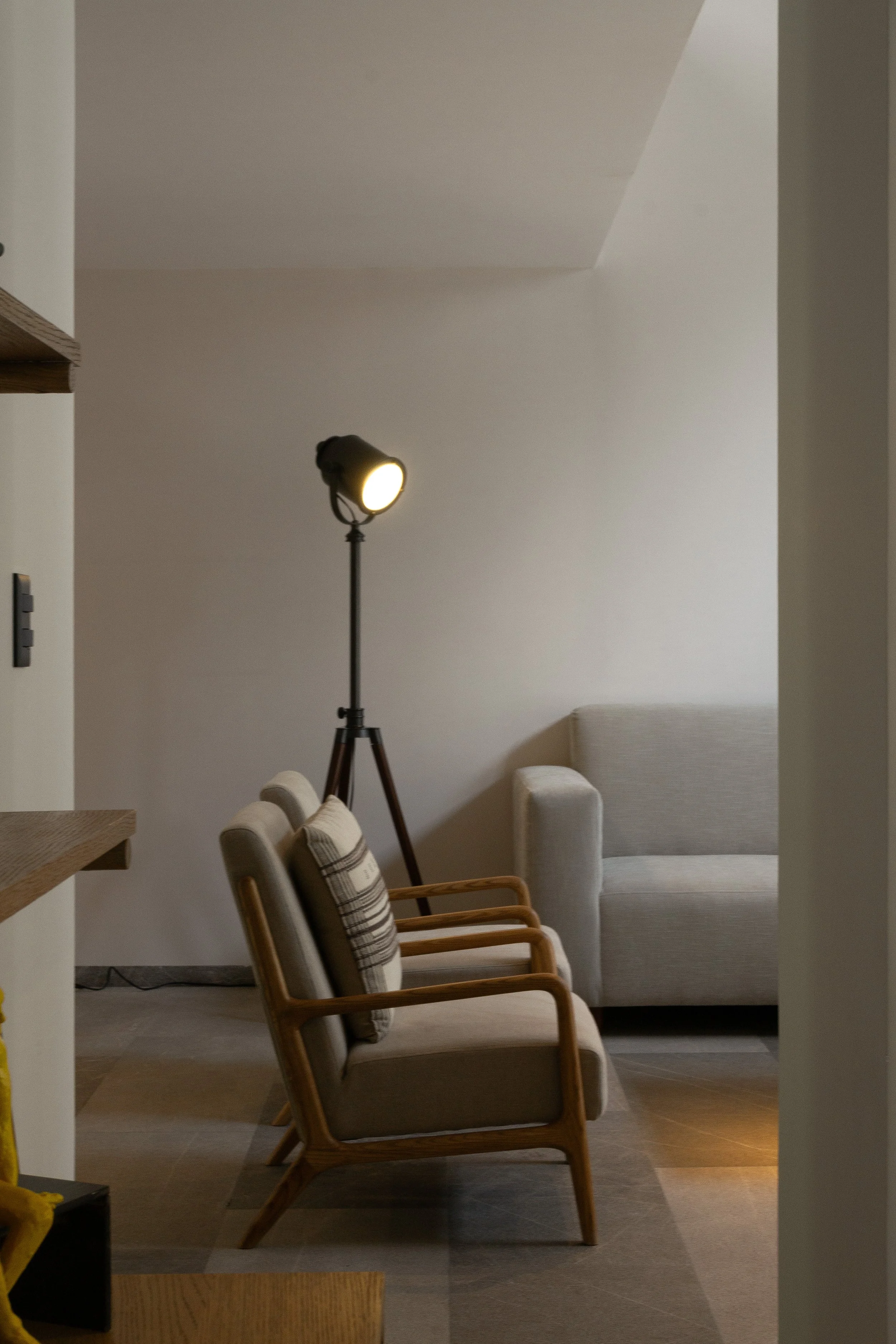 Sala de estar con sillones de madera y cojines, lámpara de pie encendida, paredes blancas y piso de madera, vista desde la entrada de la habitación.