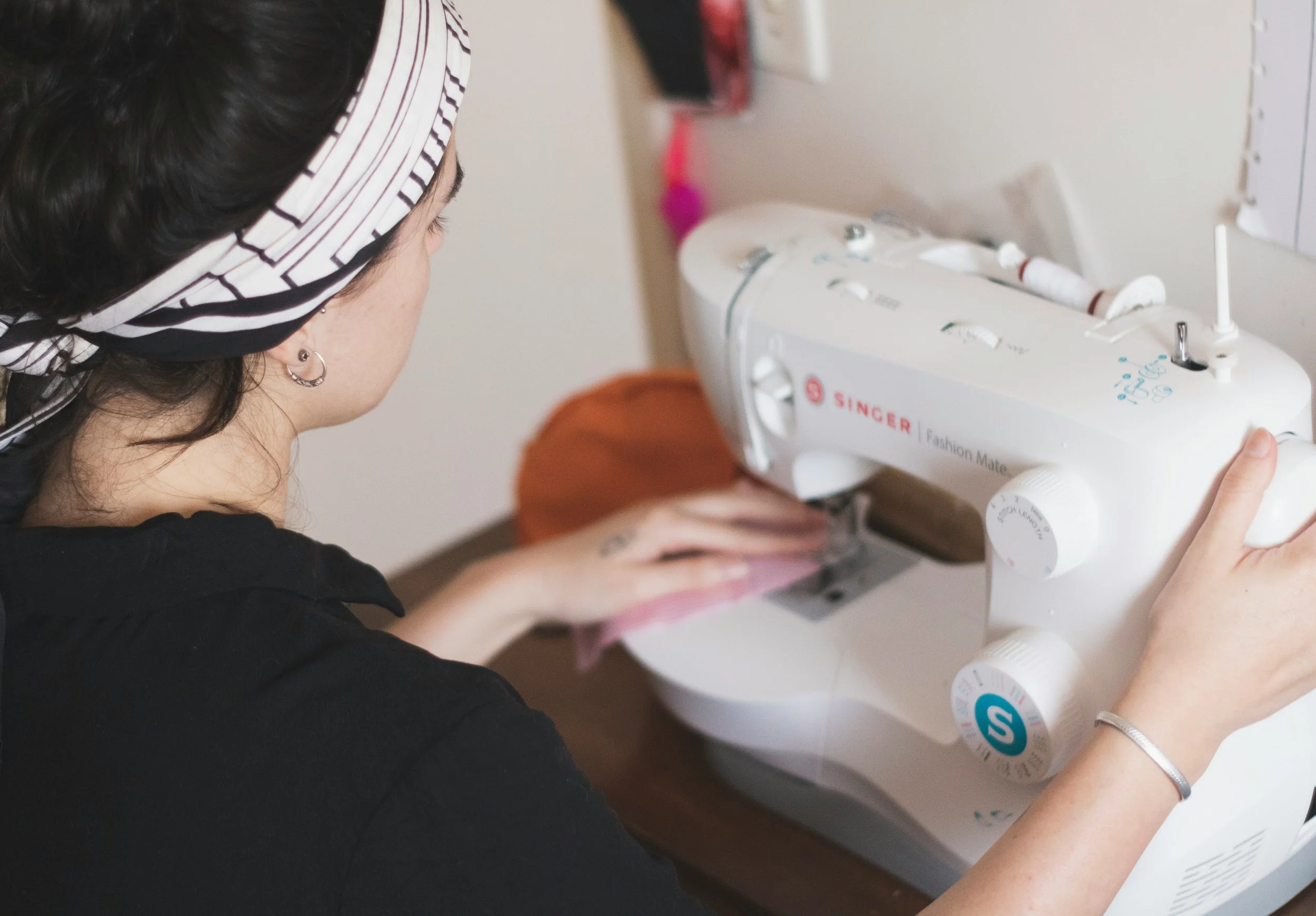 A woman sewing on a white Singer sewing machine with various buttons and dials, wearing a black shirt and a black-and-white striped headscarf.