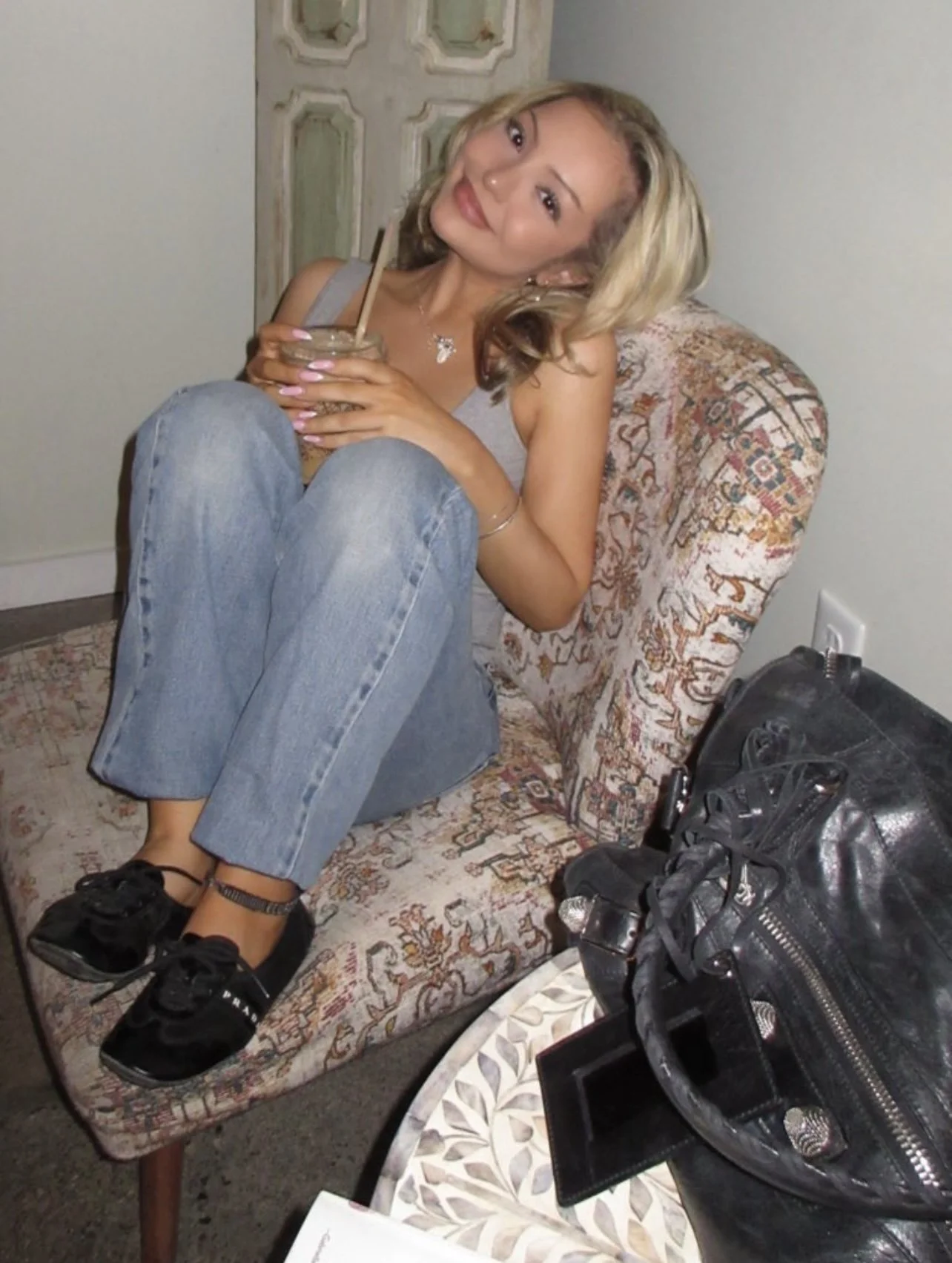 A young woman with blonde hair sitting on a patterned chair, holding a jar with a straw, smiling at the camera in a casual setting. She is wearing a tank top, jeans, and sneakers, with a necklace and bracelet. Nearby, a black bag and Instagram camera are visible on a table.