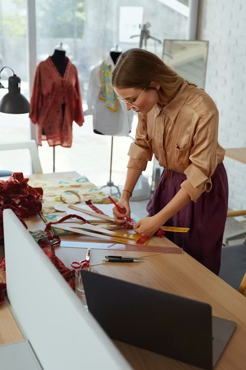 A woman in a tan blouse and purple skirt is working at a wooden table with fabric, scissors, a measuring tape, and rulers, surrounded by mannequins and fabric swatches in a bright, modern design studio.
