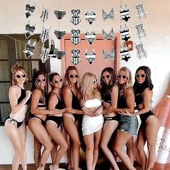 Six women in swimsuits and sunglasses standing together indoors, with butterfly decorations hanging from the ceiling above them.