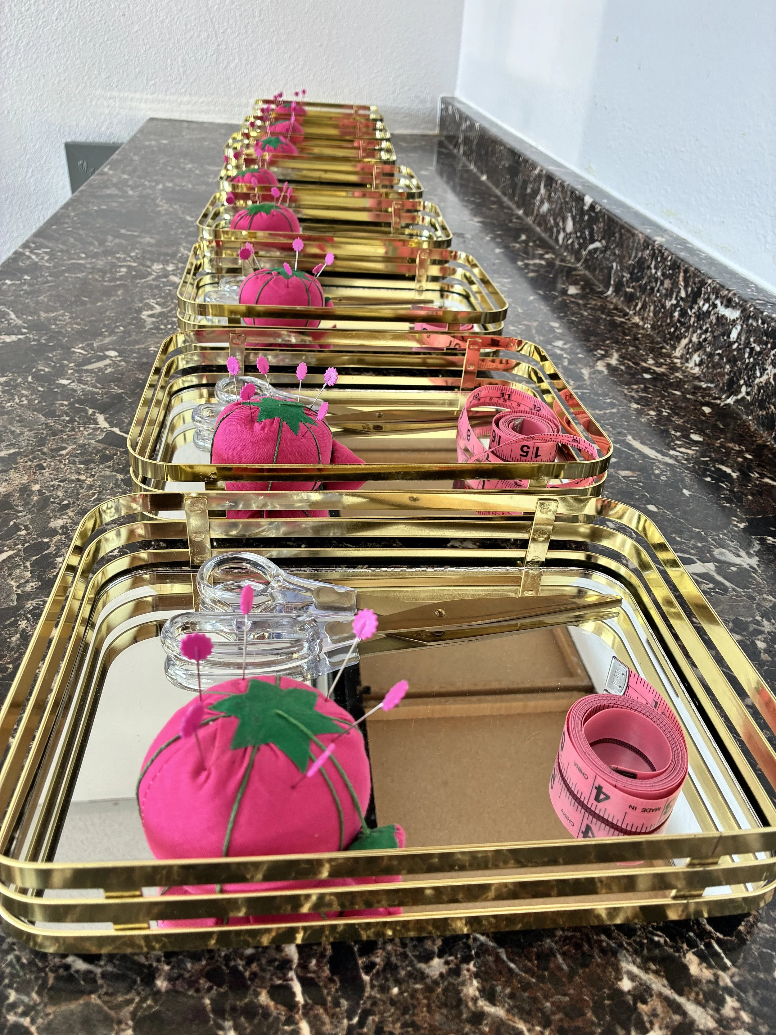 Multiple pink tomato-shaped plush pins placed on a tray with clear sharp pinheads, alongside a pink measuring tape, all arranged on a gold-colored metal rack on a dark marble countertop.