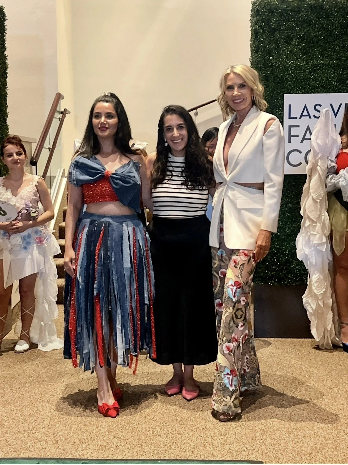 Group of women at a fashion event, dressed in colorful, stylish outfits, standing indoors with a green hedge wall and a sign in the background.