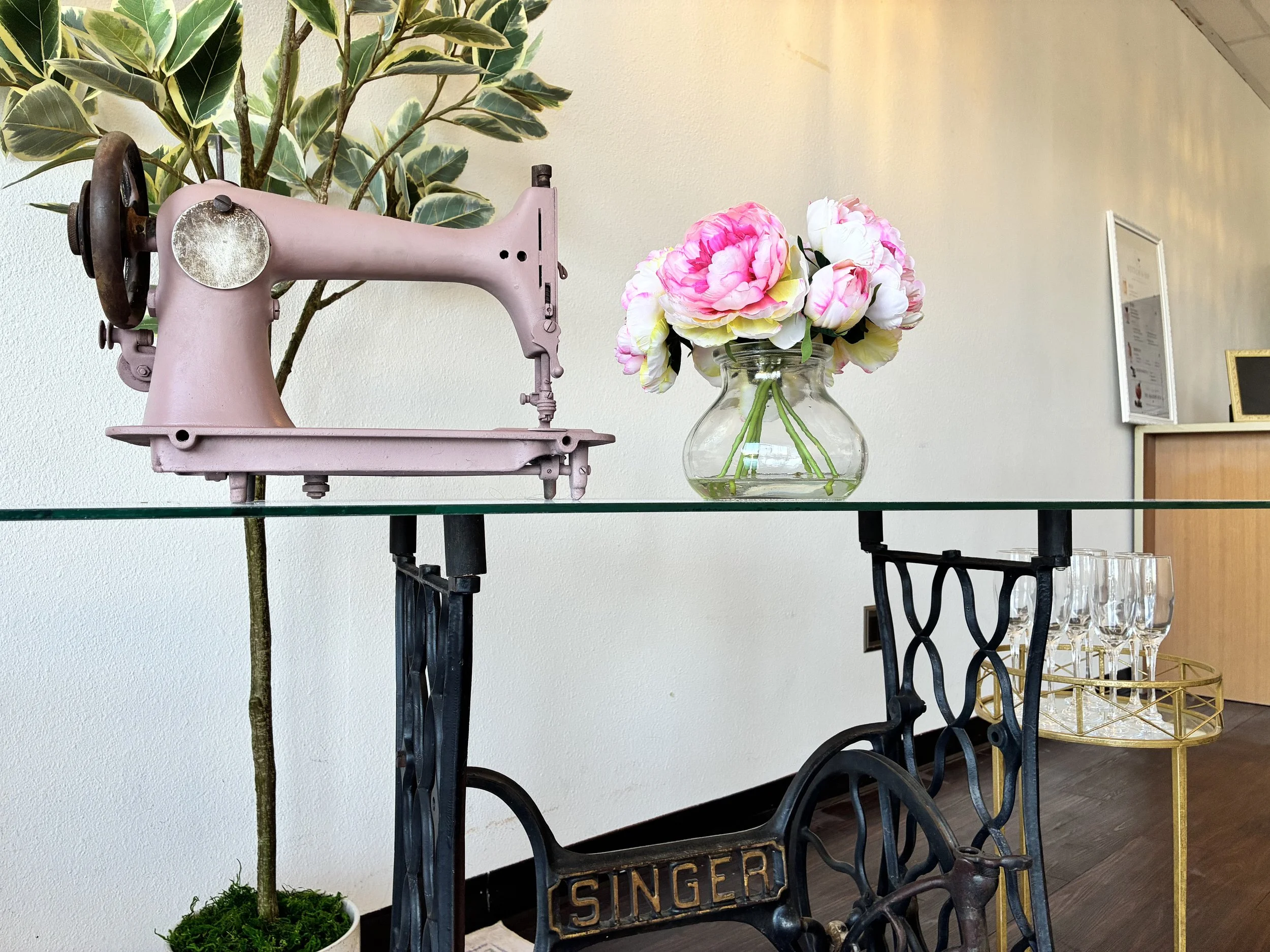 A vintage pink sewing machine on a glass table, with a bouquet of pink and white peony flowers in a glass vase, a potted plant with variegated leaves behind, and a framed certificate on the wall in the background.