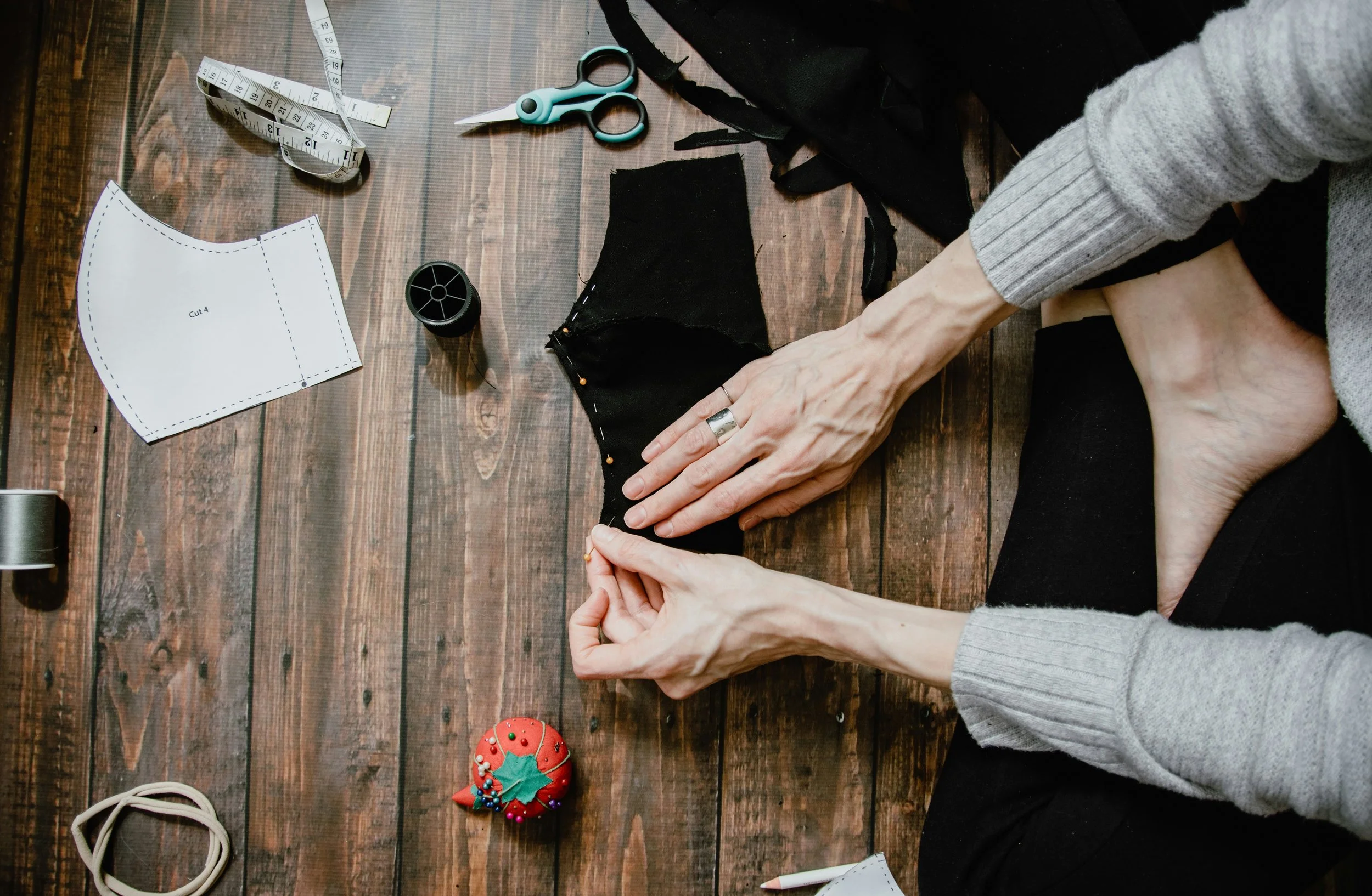 Hands working on sewing fabric on wooden table surrounded by sewing tools and accessories, such as scissors, measuring tape, thread, and a decorative pincushion.