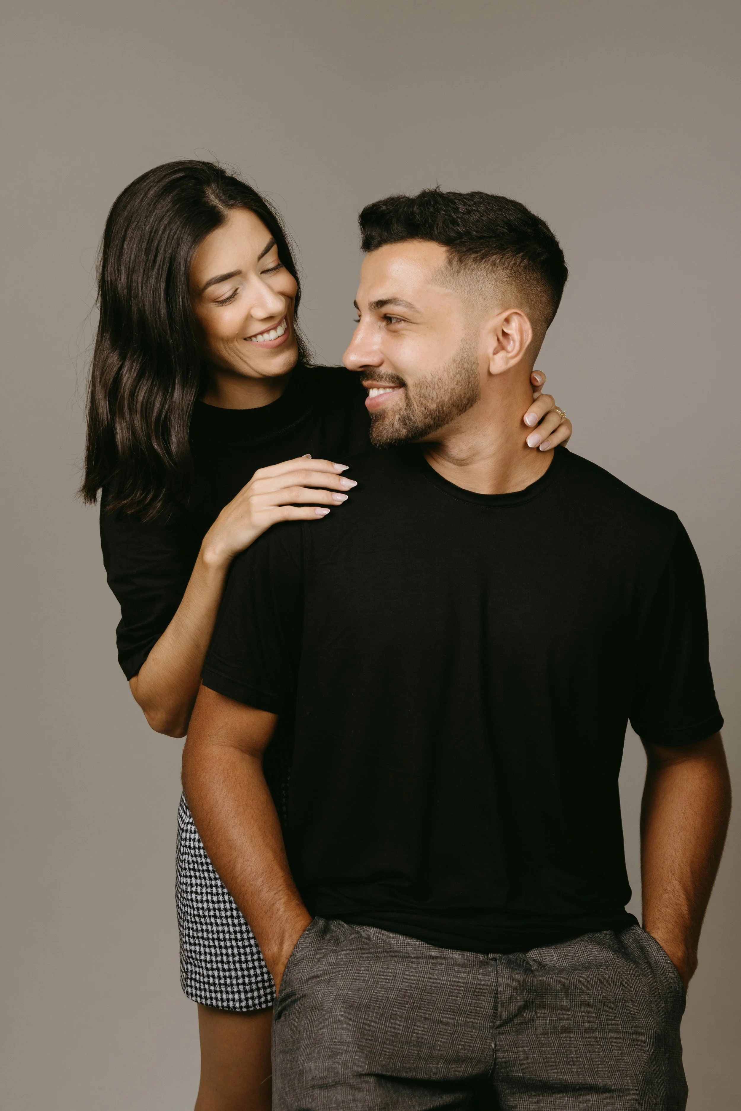 A smiling woman with long dark hair hugging and leaning close to a smiling man with short dark hair, both dressed in black, standing against a plain gray background.