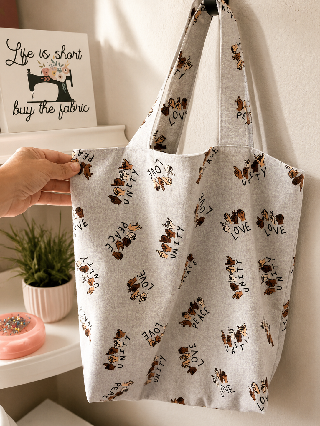 A tote bag with a pattern of hands making peace, love, and unity signs, hanging on a wall.