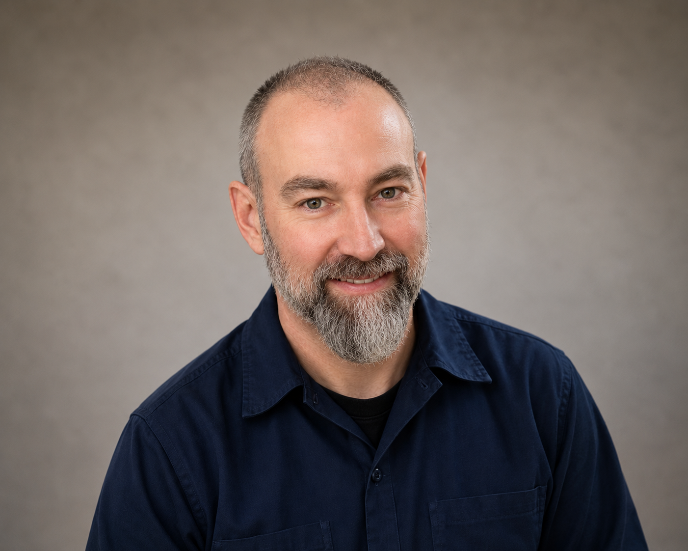 Headshot of a middle-aged man with a beard and short hair, wearing a dark blue shirt, smiling against a neutral gray background.
