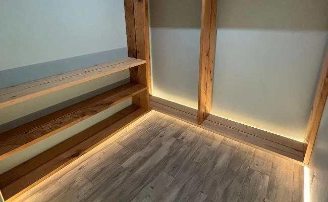 Corner of a room with wooden shelves and baseboards, with LED strip lighting installed under the shelves and along the base of the wall.