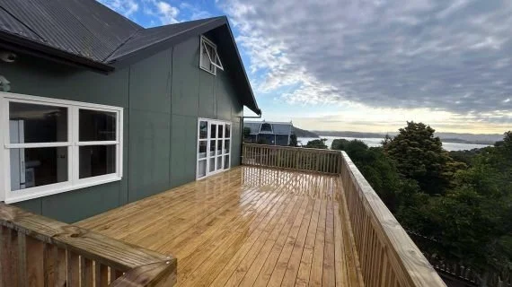 Large wooden deck attached to a green house with white-framed windows, overlooking a landscape of trees and water under a cloudy sky.