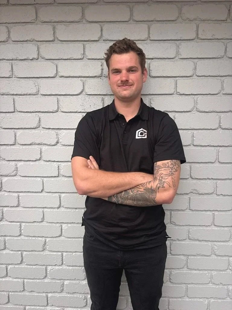 A young man with light skin, short curly hair, and a slight beard, standing with crossed arms, in front of a white brick wall. He's wearing a black polo shirt with a logo on the chest and black pants, with visible tattoos on his left arm.