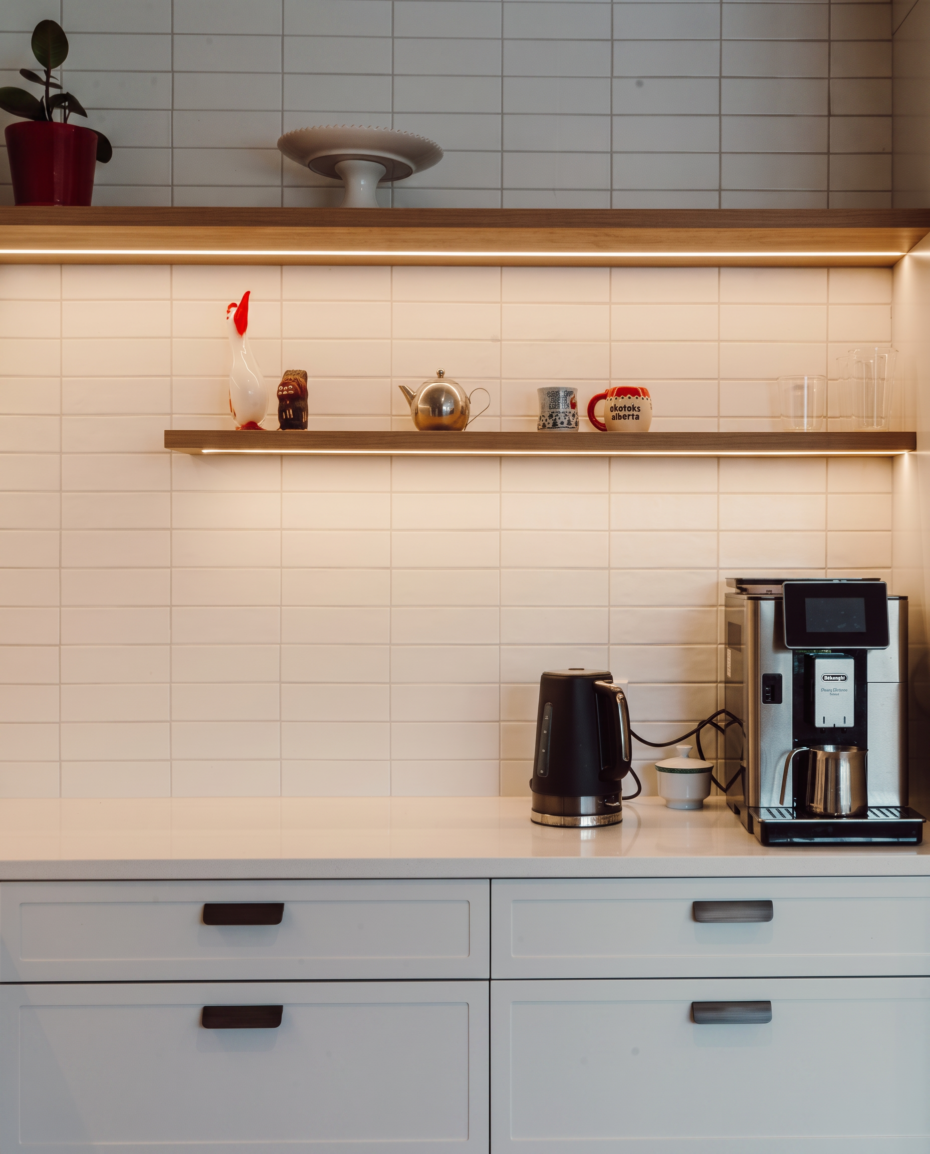 White kitchen countertop with black kettle, coffee machine, and two mugs. Overhead shelves with decorative items, including a red potted plant, ceramic rooster, teapot, and glass containers, against a white tiled wall.