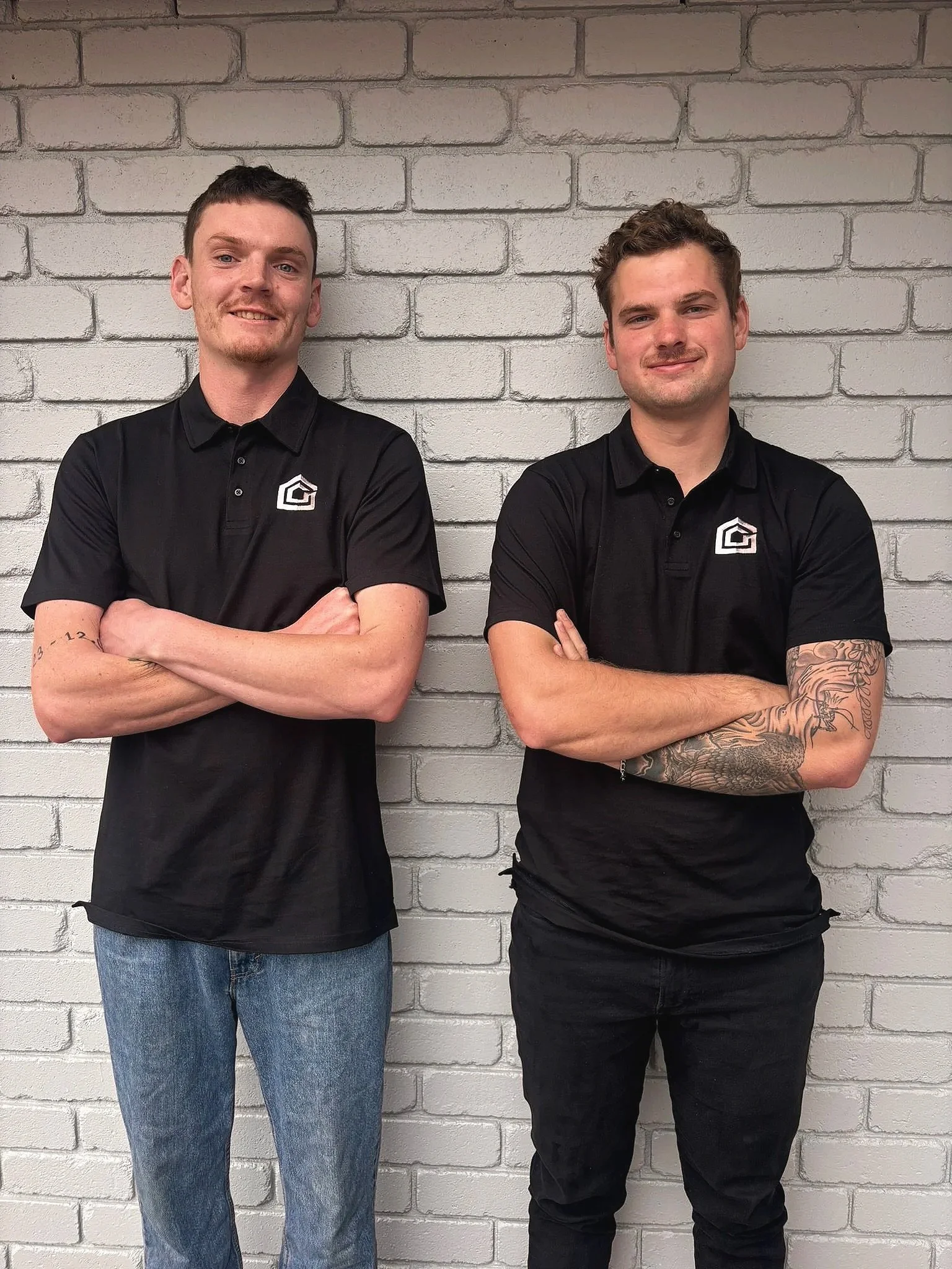 Two young men standing side by side in front of a white brick wall with their arms crossed, wearing black polo shirts with a logo on the chest.