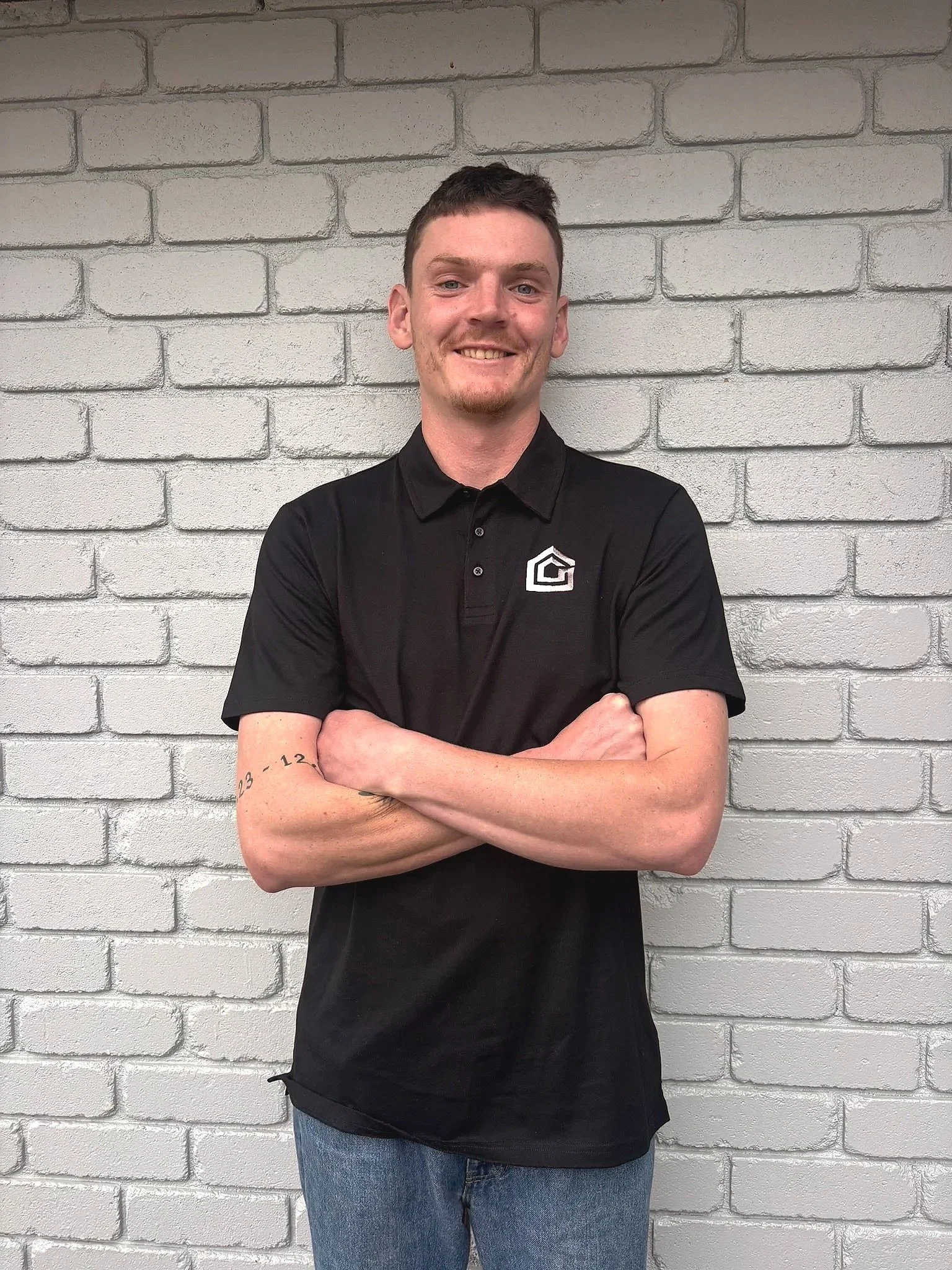 A young man with short dark hair and a small goatee smiling, standing with his arms crossed in front of a white brick wall. He is wearing a black polo shirt with a logo on the chest and blue jeans.
