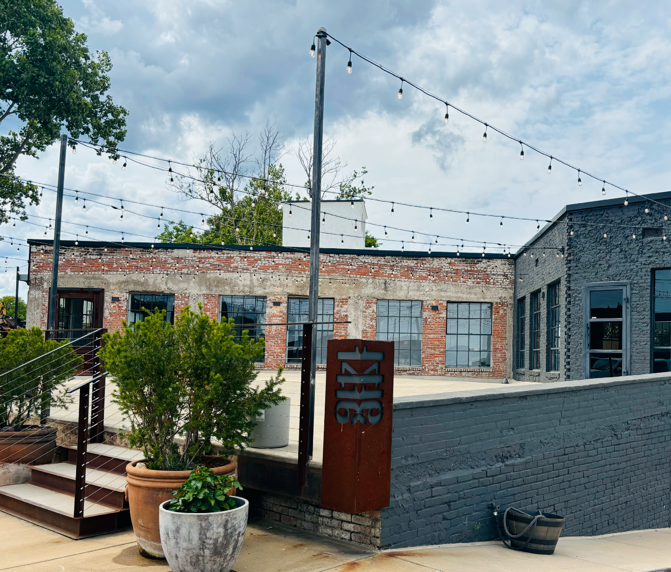 Outdoor rooftop patio with string lights, potted plants, and aged brick building with large windows under a partly cloudy sky.